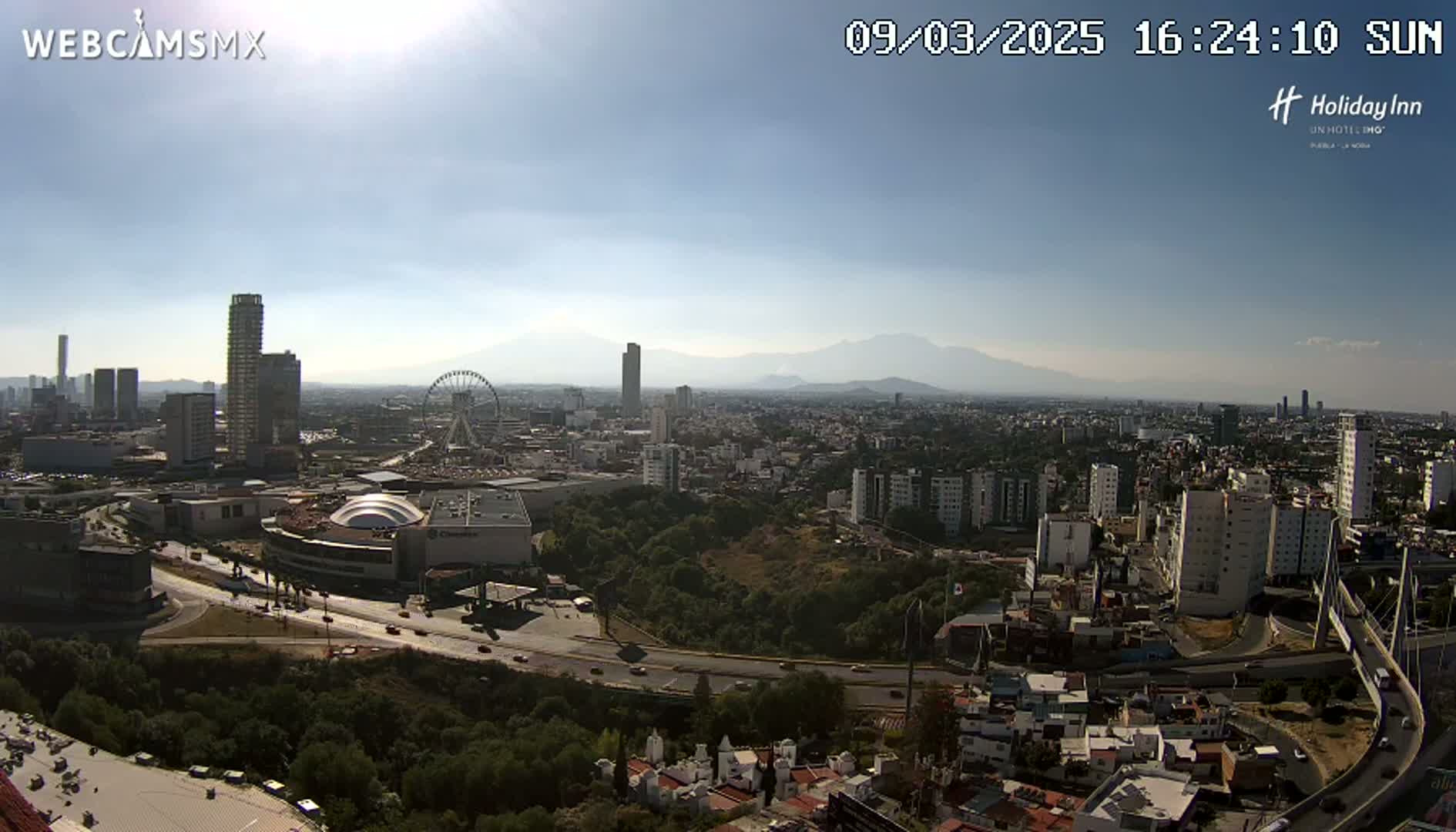 A sunny, high-angle view shows a sprawling city with a Ferris wheel, skyscrapers, and mountains in the distance.