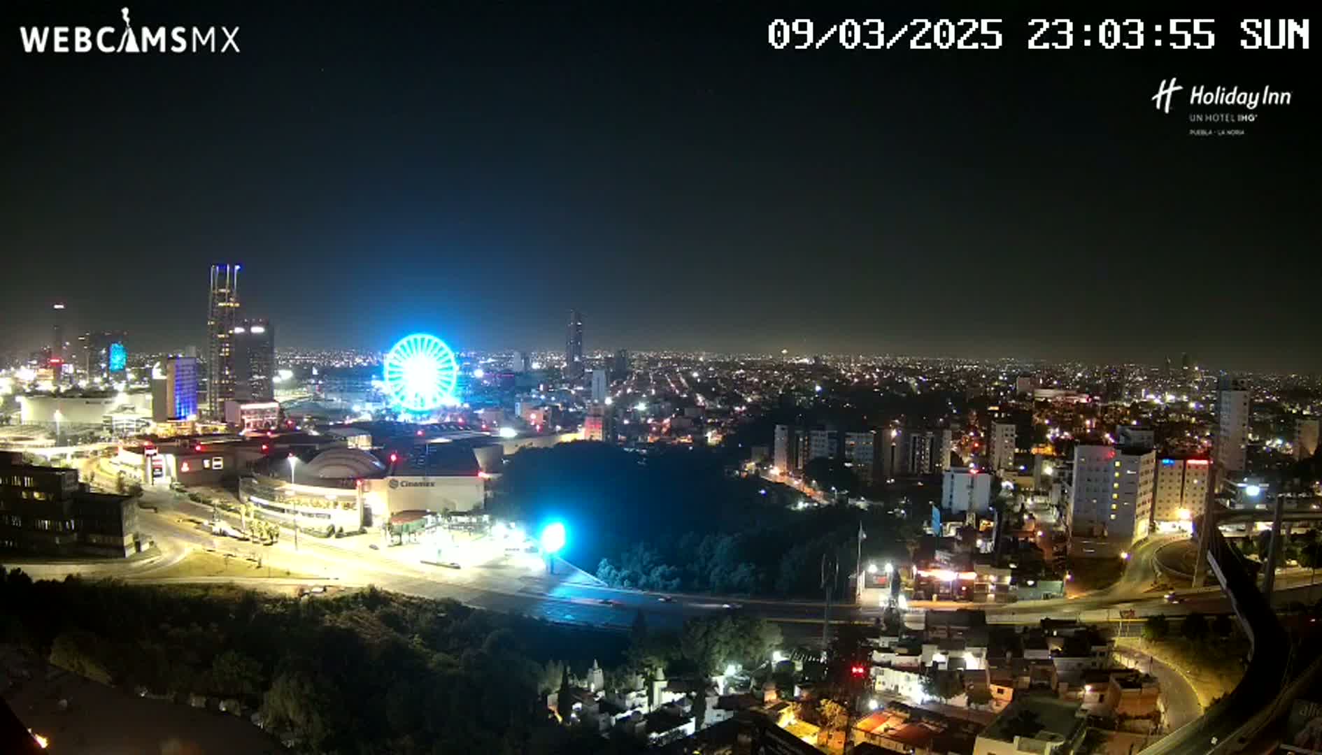 A nighttime cityscape with a Ferris wheel, illuminated buildings, and roads, under clear skies.