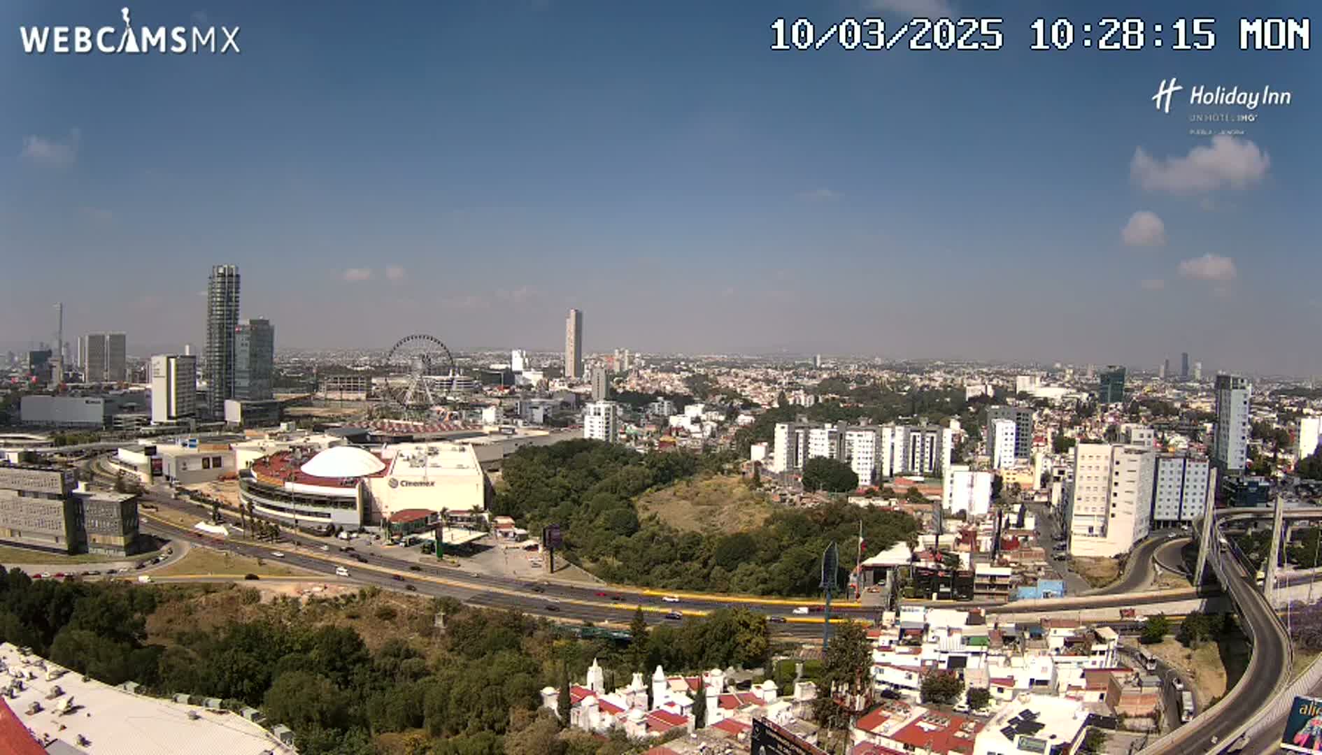 A sprawling cityscape under a clear, sunny sky, featuring a Ferris wheel, several high-rise buildings, and a large highway system.
