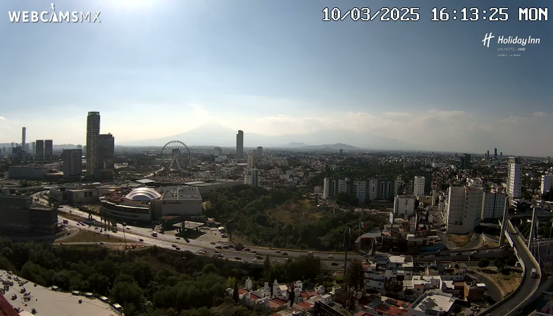 A sprawling city landscape under a mostly sunny sky, featuring high-rise buildings, a Ferris wheel, and a large highway system, with mountains visible in the distance.