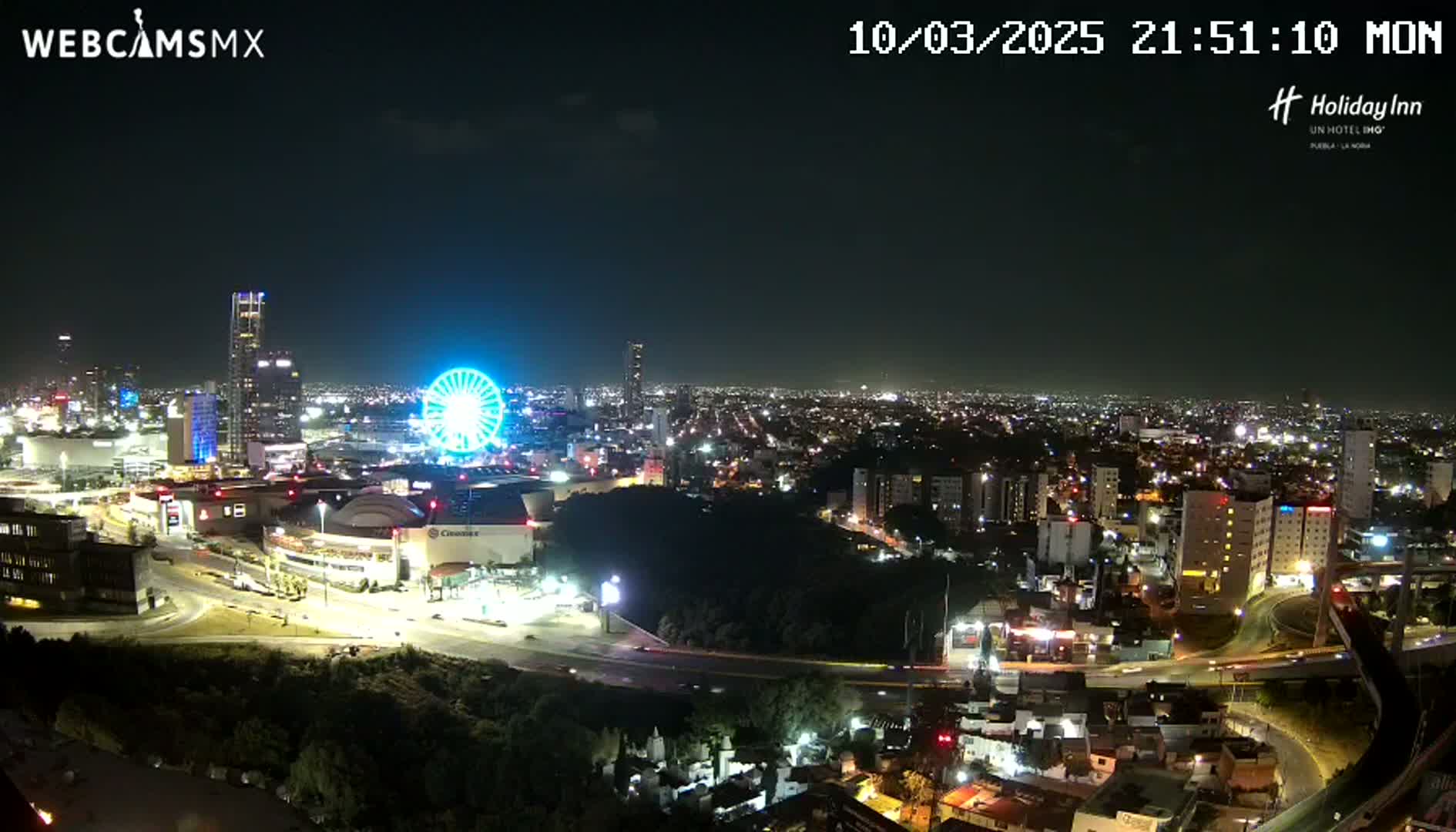 A nighttime aerial view of a city illuminated by lights, featuring a Ferris wheel and several tall buildings, under a clear sky.