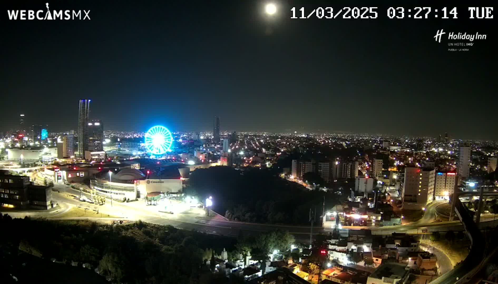 A brightly lit city at night, featuring a Ferris wheel and a full moon in a clear sky, is seen from an elevated vantage point.