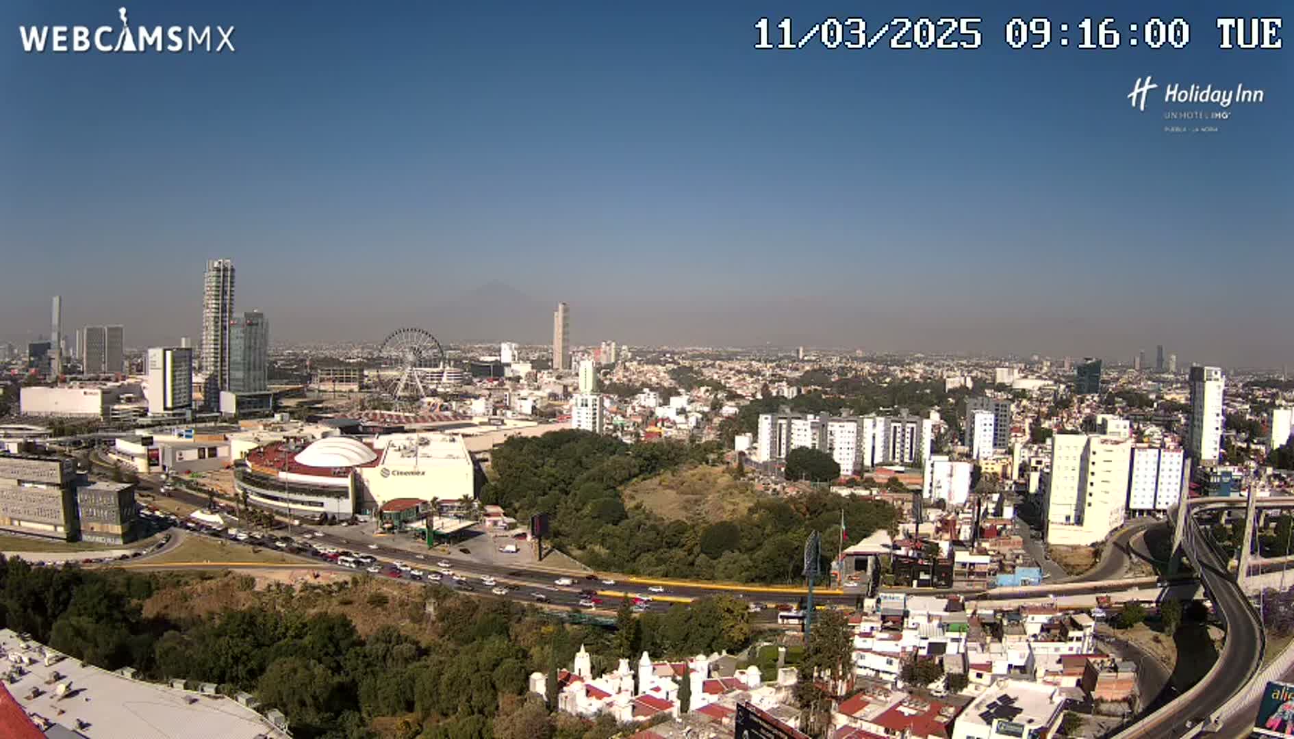 A sprawling city landscape under a clear blue sky shows a mix of high-rise buildings, a Ferris wheel, and extensive roadways with busy traffic.
