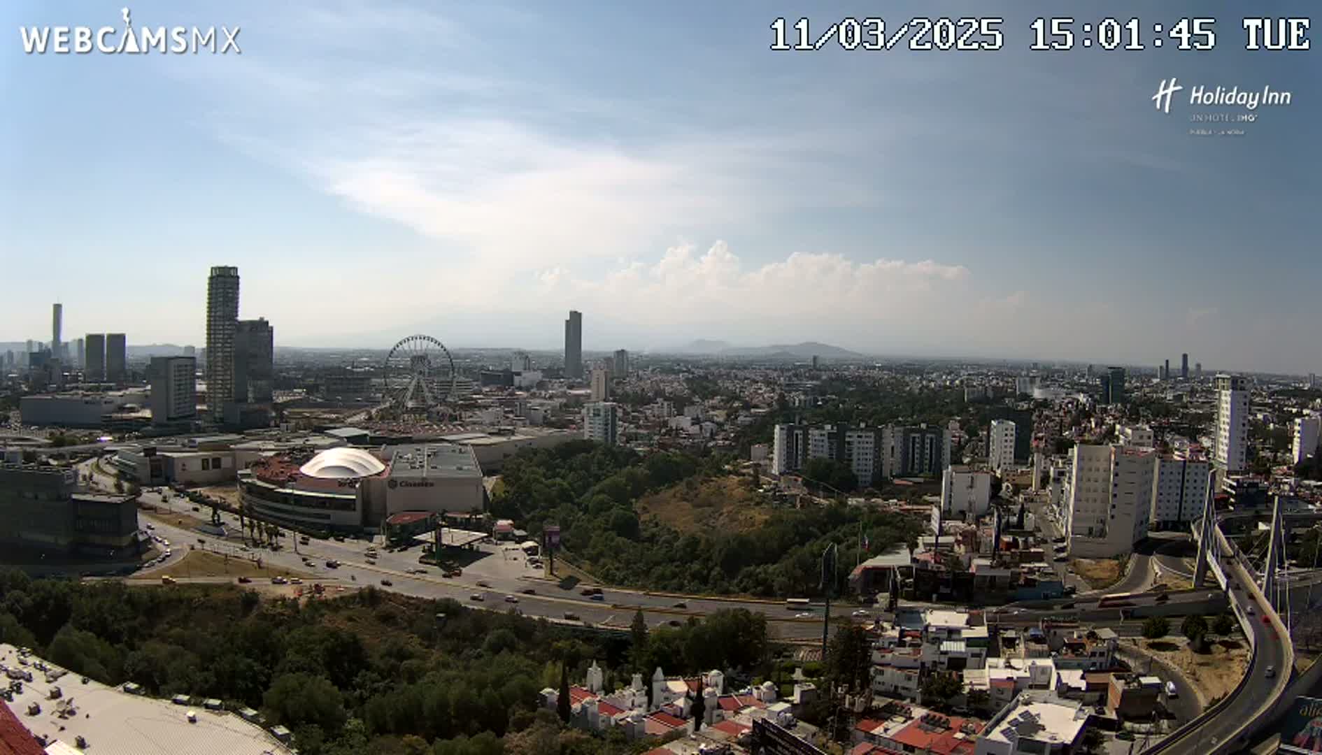 A sprawling city landscape under a partly cloudy sky shows a Ferris wheel, several high-rise buildings, and a large shopping mall, with a highway curving through the scene.