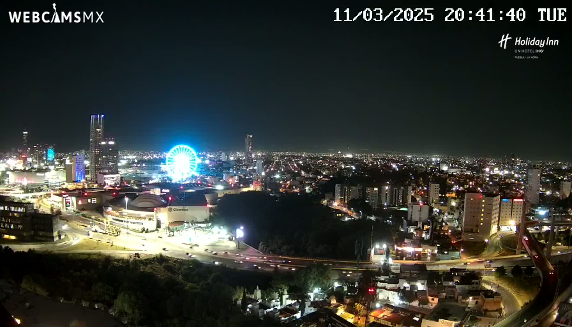 A nighttime, high-angle view of a city featuring a brightly lit Ferris wheel near a large shopping mall and a highway system weaving through the urban landscape.
