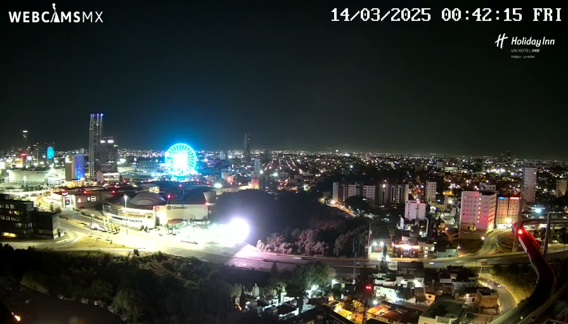 A nighttime aerial view of a city illuminated by lights, featuring a Ferris wheel and a large stadium, under a clear sky.