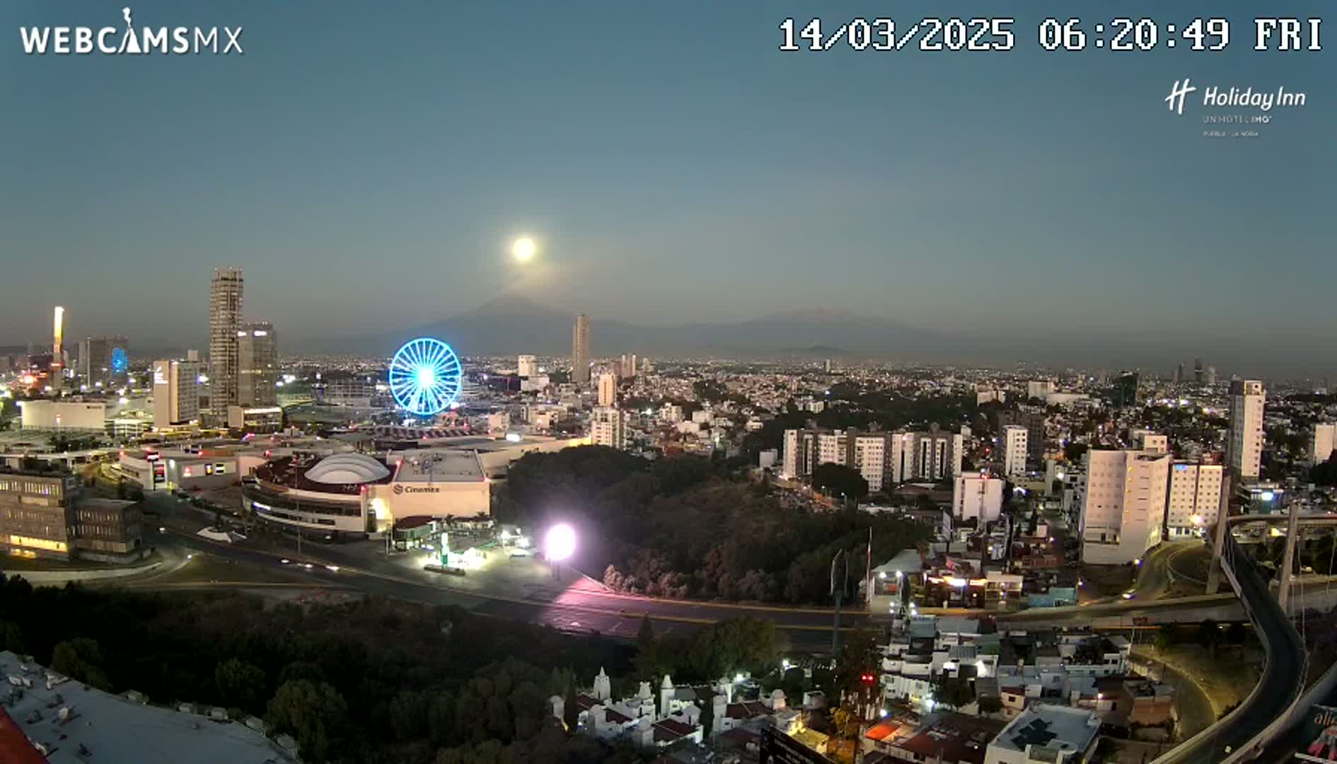 A nighttime cityscape with a Ferris wheel, illuminated buildings, and a distant mountain range under a clear, moonlit sky.
