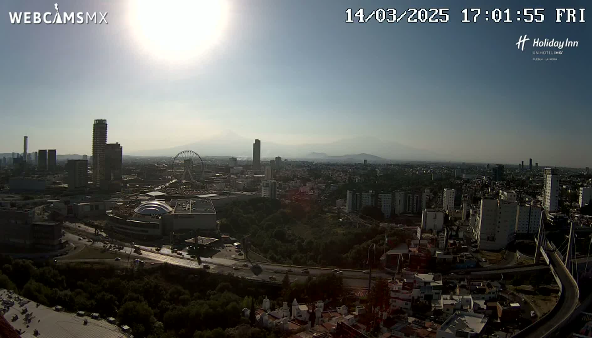 A sunny, hazy day reveals a sprawling cityscape with a Ferris wheel, numerous buildings, and a highway system, all framed by distant mountains.