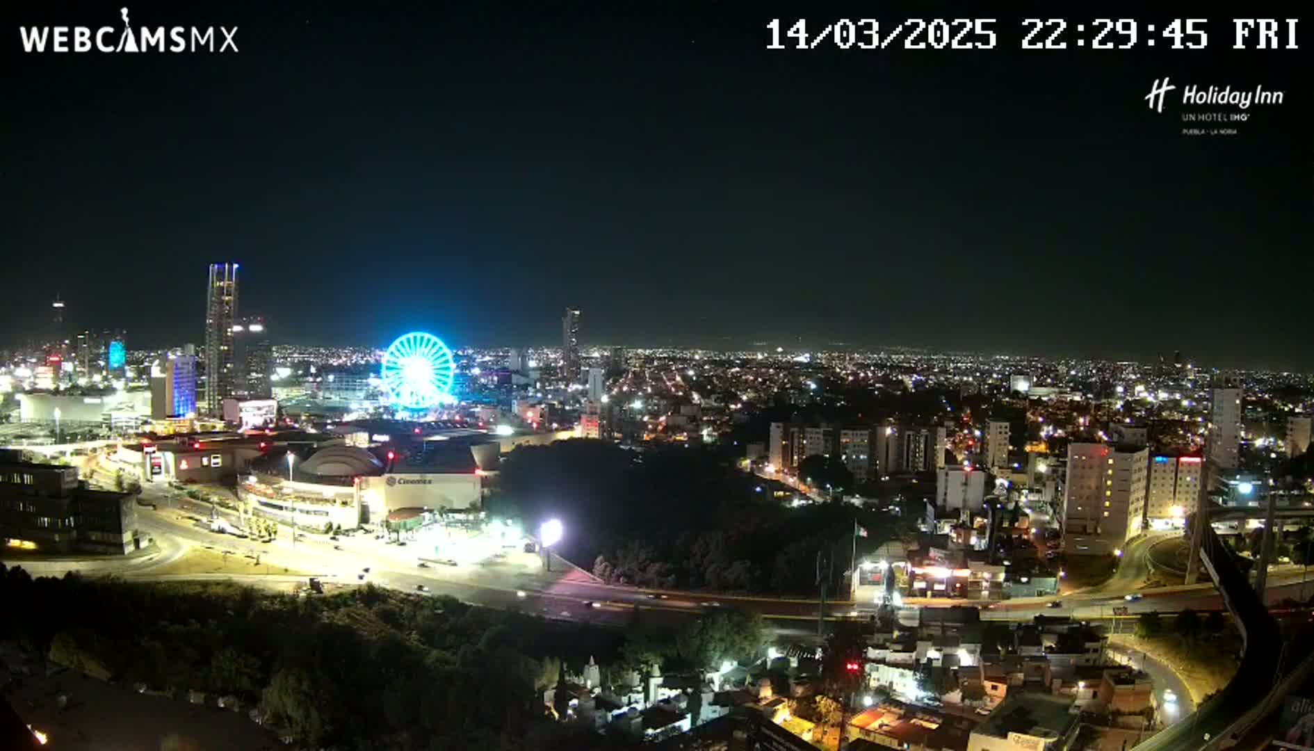 A nighttime aerial view of a city illuminated with many lights, featuring a Ferris wheel and a large shopping center, under a clear sky.