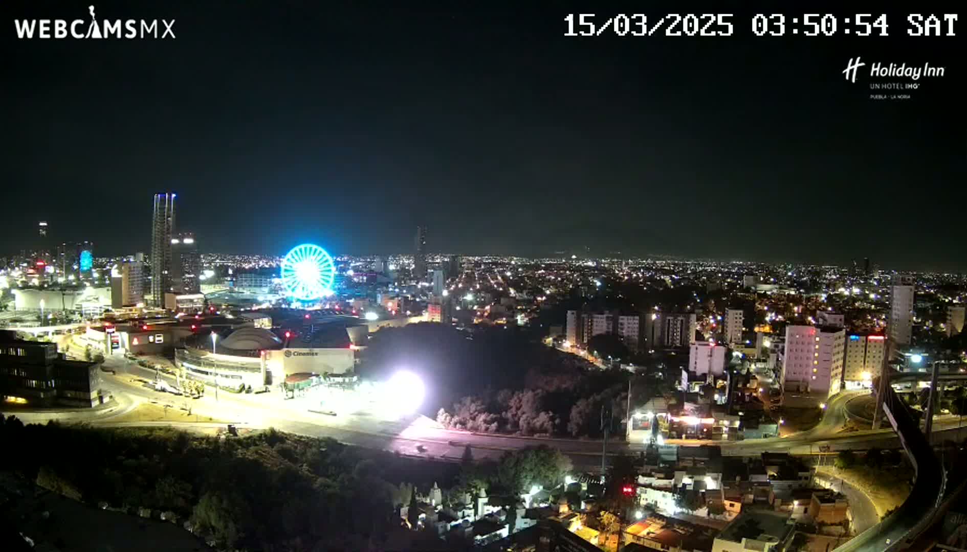 A nighttime, clear-weather view of a city illuminated by lights, featuring a Ferris wheel and several buildings, including a large stadium or arena.