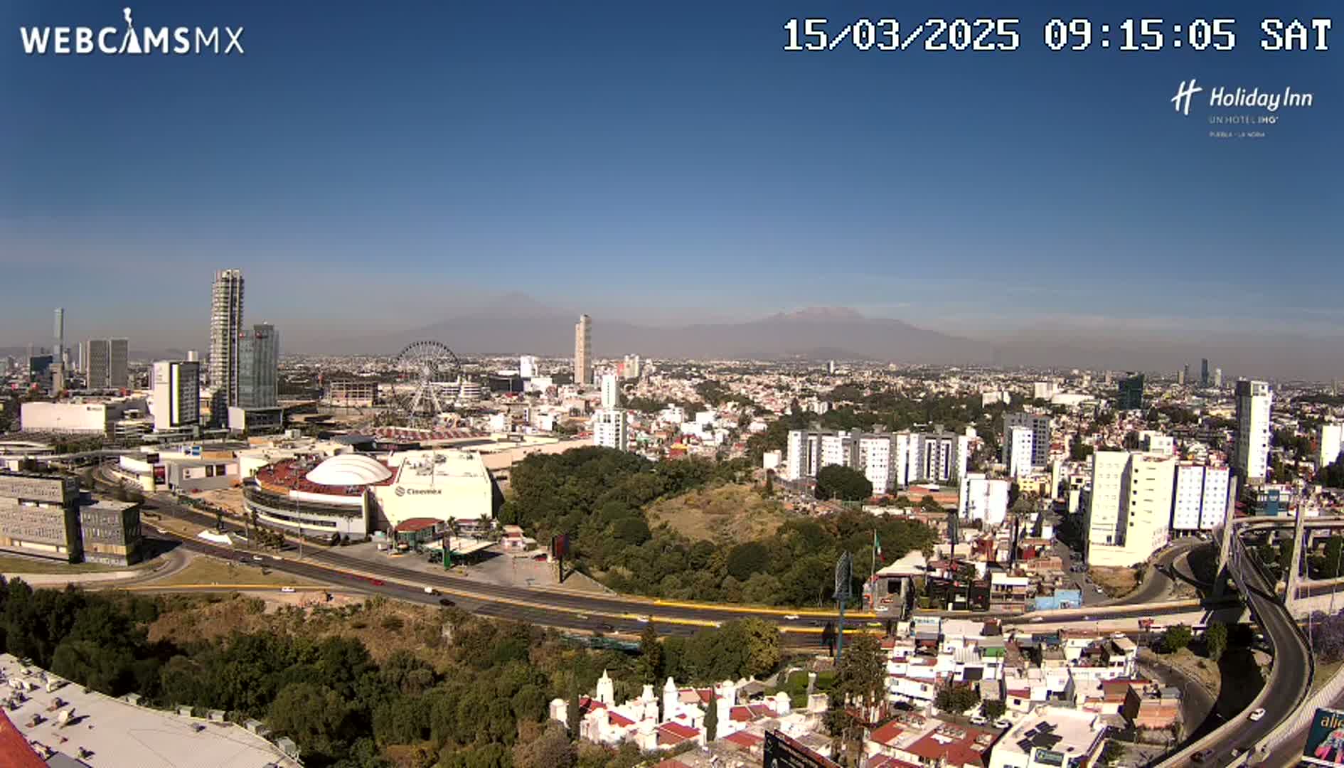 A sprawling city with high-rise buildings, a Ferris wheel, and a large dome-shaped structure is visible under a clear, sunny sky, with mountains in the distance.