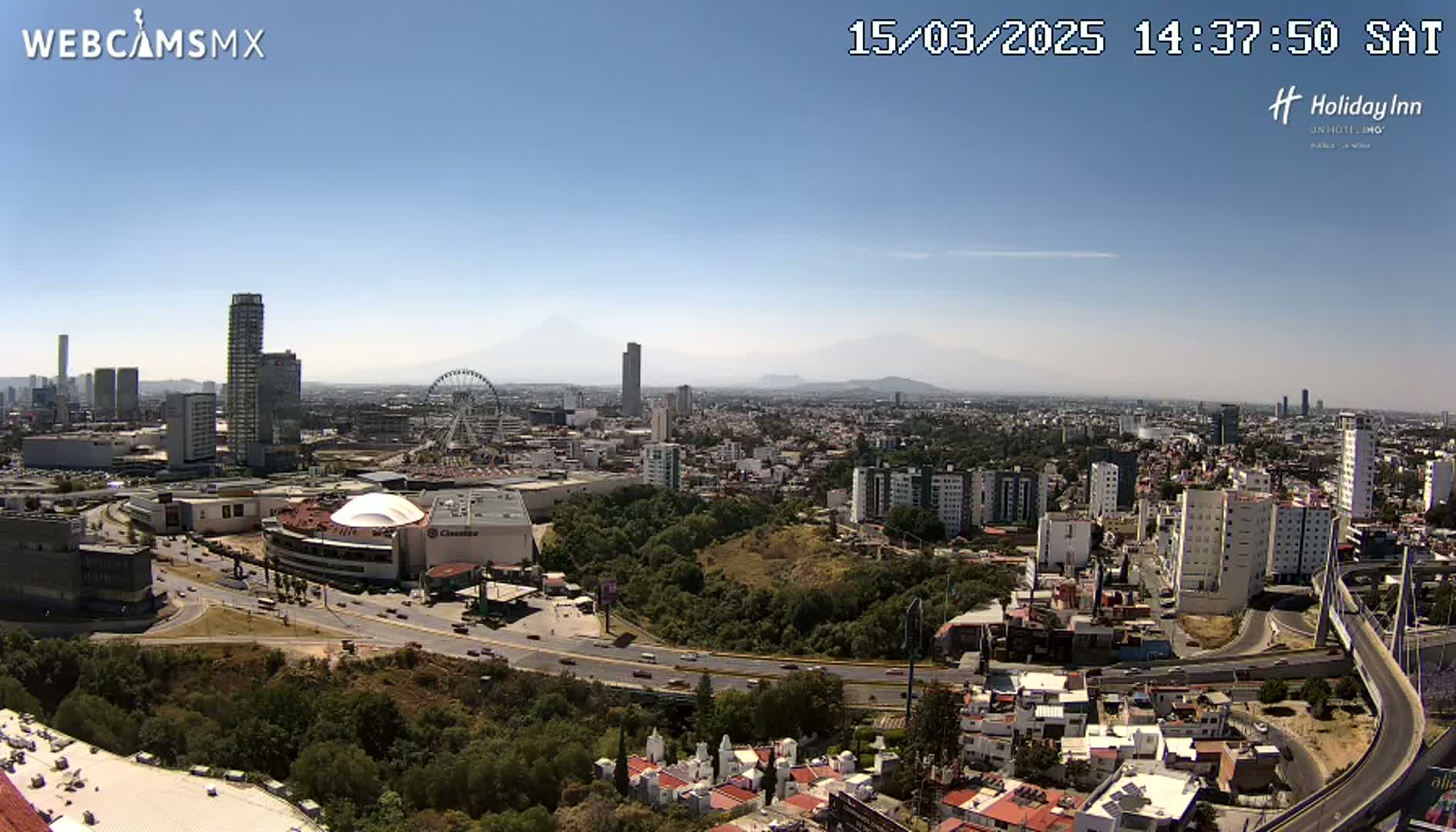 A high-angle, long shot on a clear, sunny day shows a sprawling city with numerous buildings, a Ferris wheel, and mountains in the distance.
