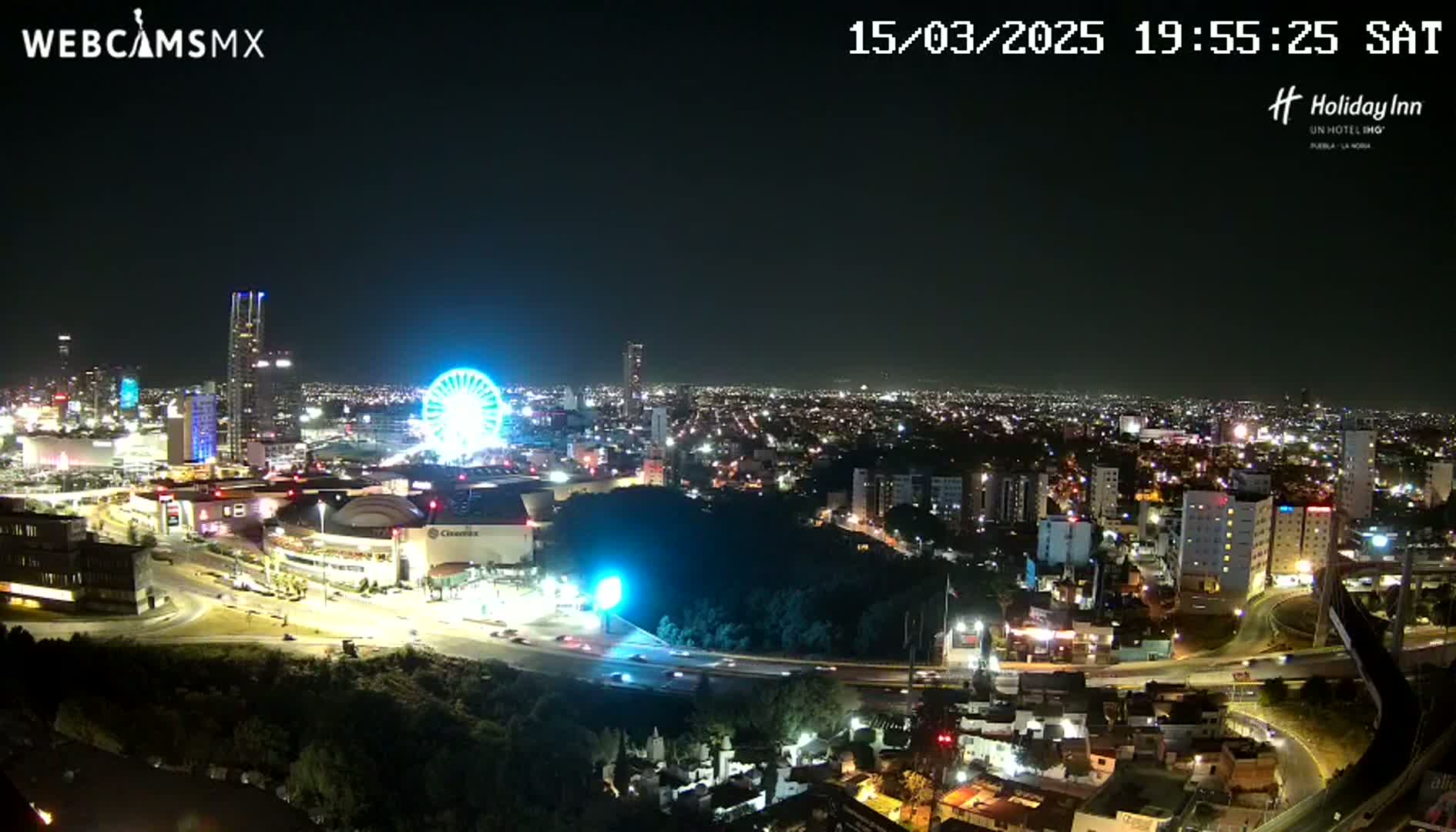 A nighttime aerial view of a city with a brightly lit Ferris wheel and numerous buildings, under clear skies.