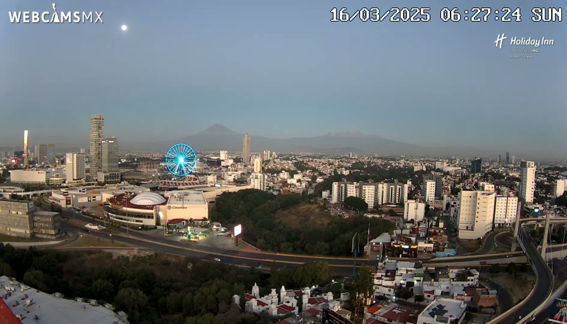 A clear sunrise view of a city with a Ferris wheel, mountains in the distance, and a highway system winding through the urban landscape.