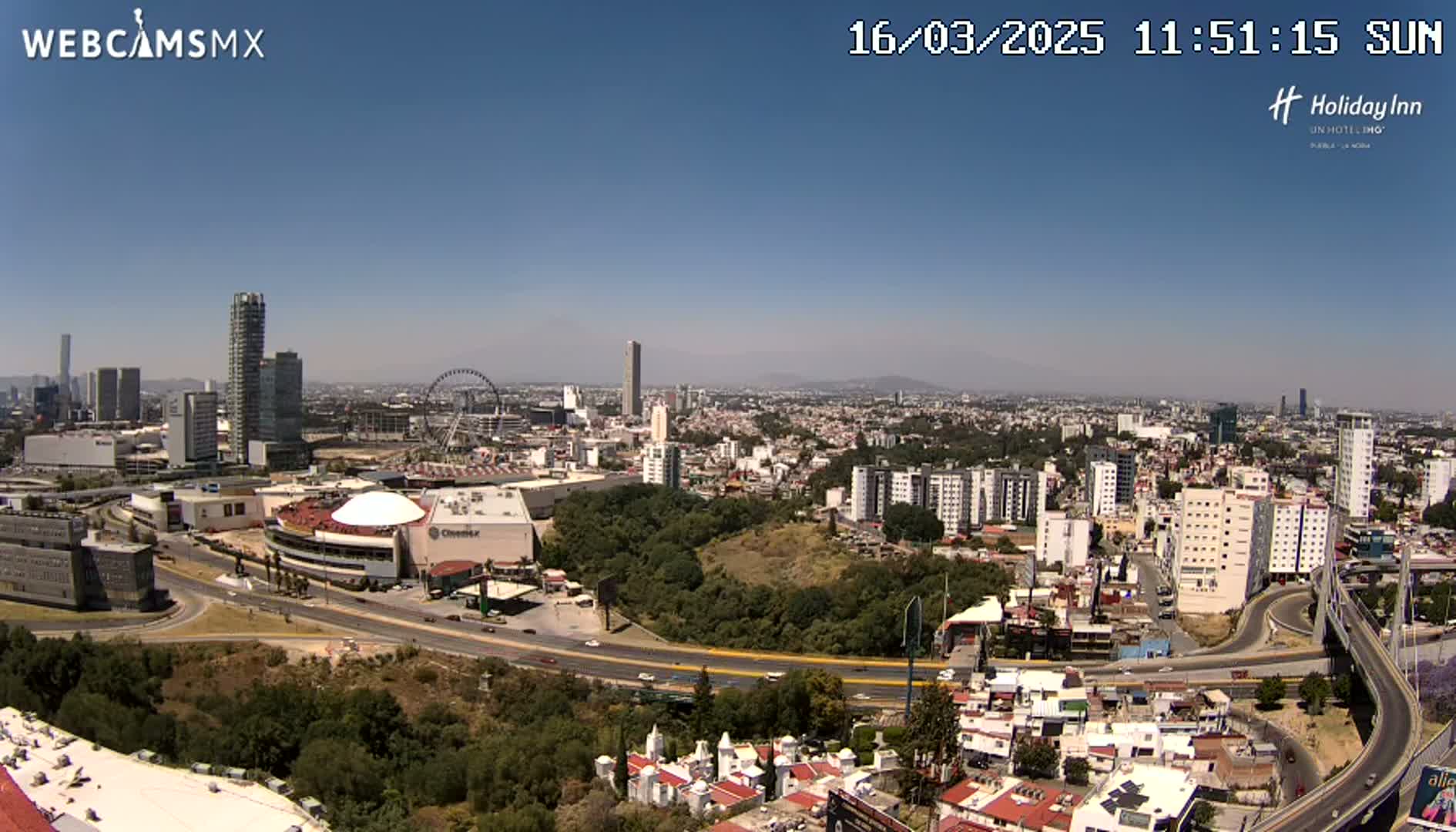 A sprawling city landscape under a clear blue sky shows a mix of high-rise buildings, residential areas, a Ferris wheel, and a large highway curving through the scene.