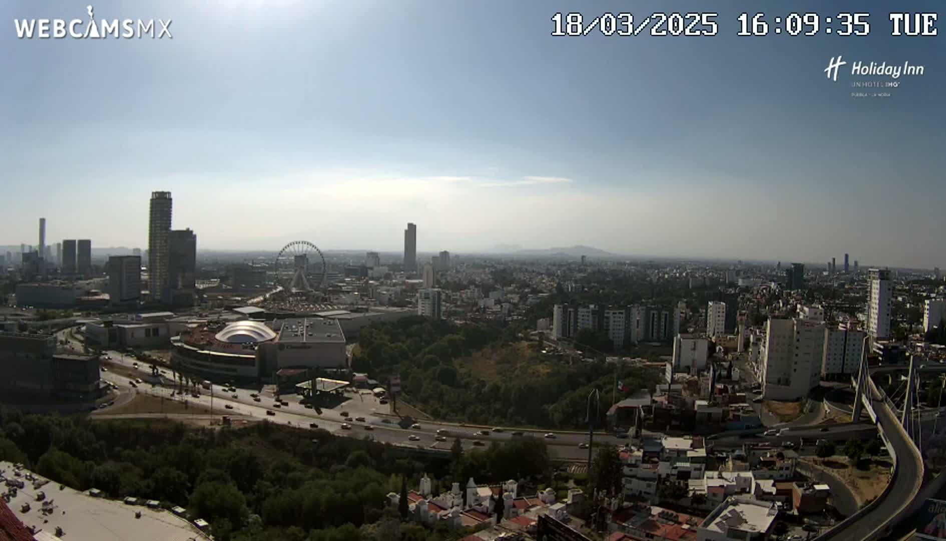 A sunny, hazy day overlooks a sprawling city with numerous high-rise buildings, a Ferris wheel, a large shopping mall, and a highway system, all surrounded by green hills and vegetation.