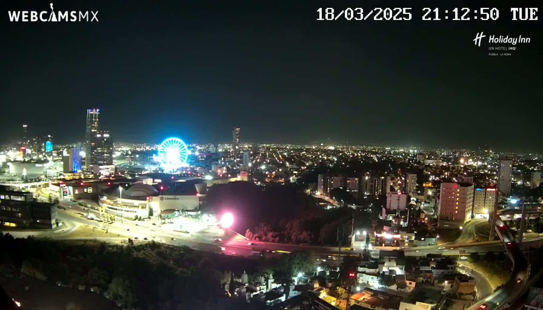 A nighttime, aerial view of a city with a Ferris wheel illuminated, surrounded by brightly lit buildings and roads.