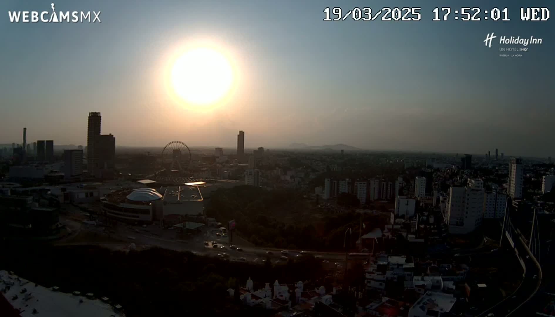 A hazy sunset illuminates a sprawling city landscape featuring skyscrapers, a Ferris wheel, and a large shopping mall.