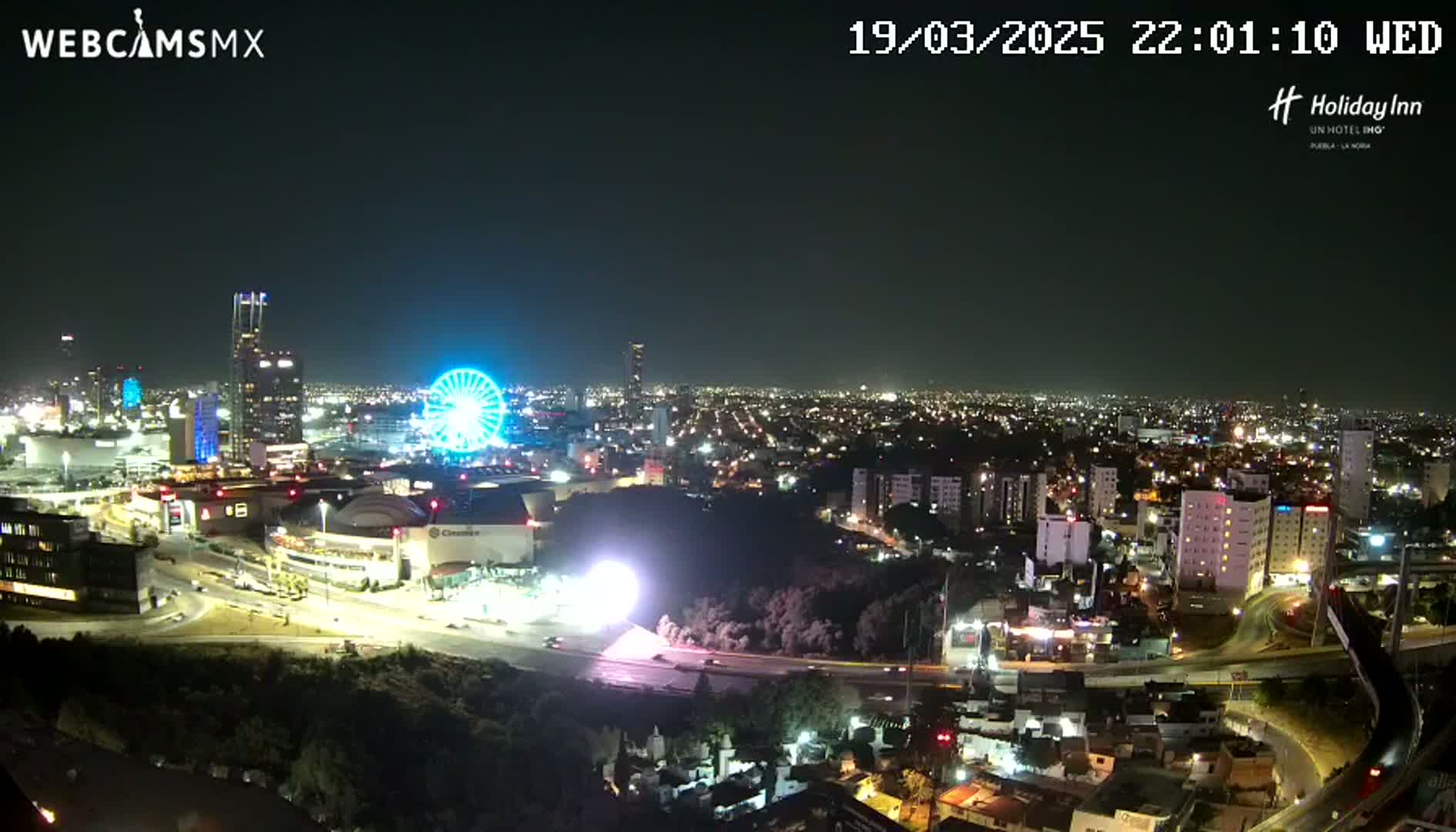 A nighttime aerial view of a city with a Ferris wheel and illuminated buildings, under a clear sky.