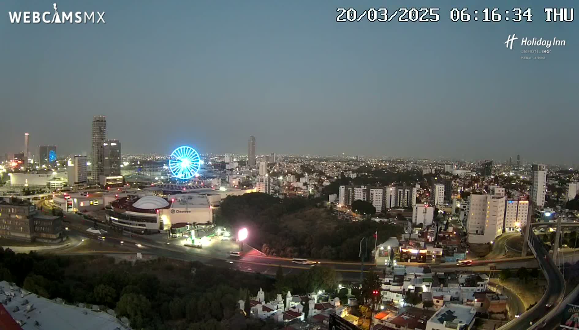 A nighttime, high-angle view of a city illuminated with lights, featuring a Ferris wheel, several buildings, and a highway system under a twilight sky.