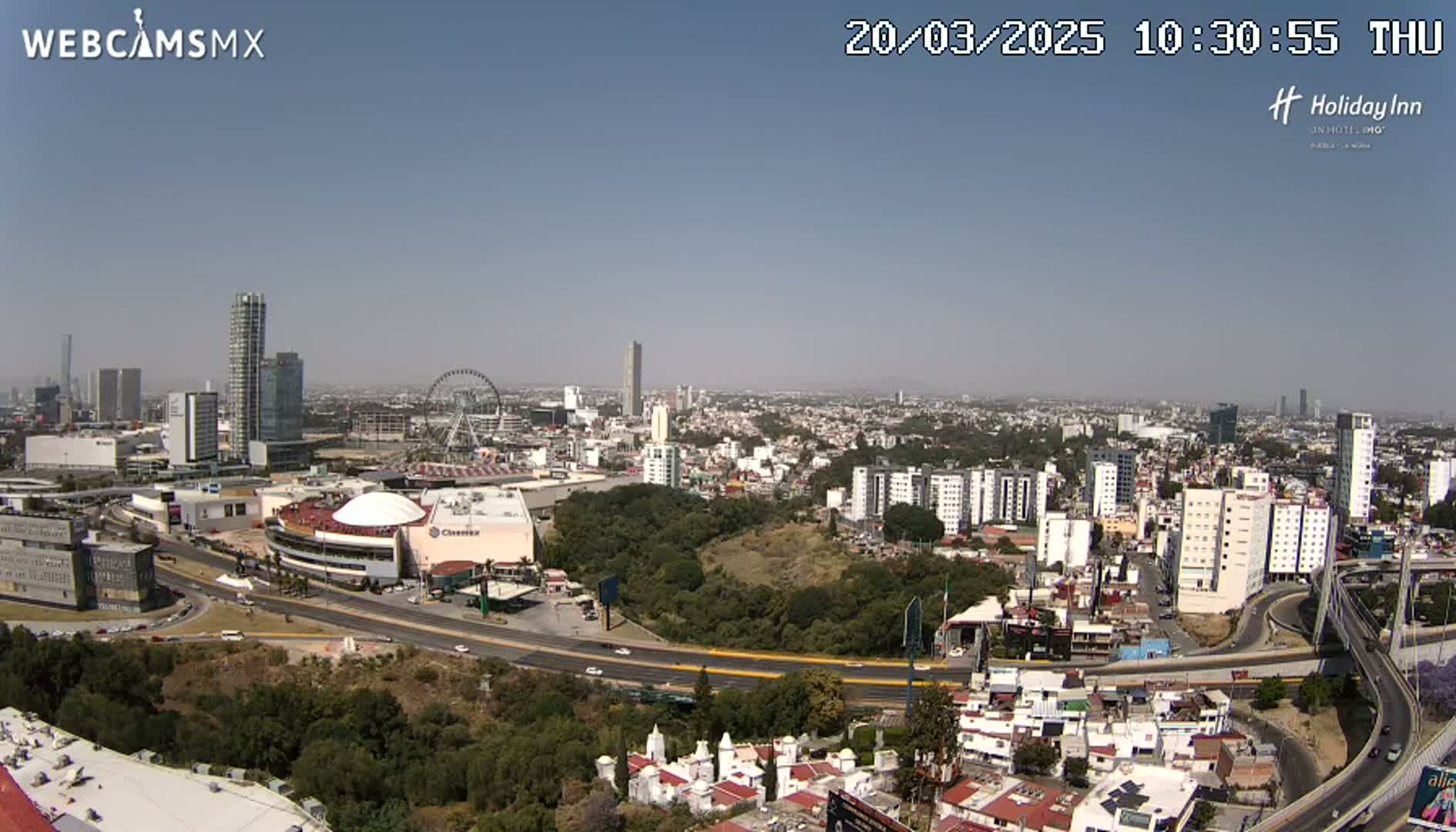 A sprawling cityscape under a clear blue sky, featuring high-rise buildings, a Ferris wheel, and a large roadway system winding through a green area.