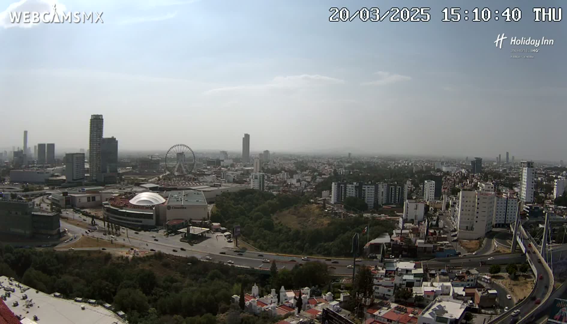 A hazy, sunny day reveals a sprawling cityscape with high-rise buildings, a Ferris wheel, and a large shopping mall, all situated near a major highway and bridge.