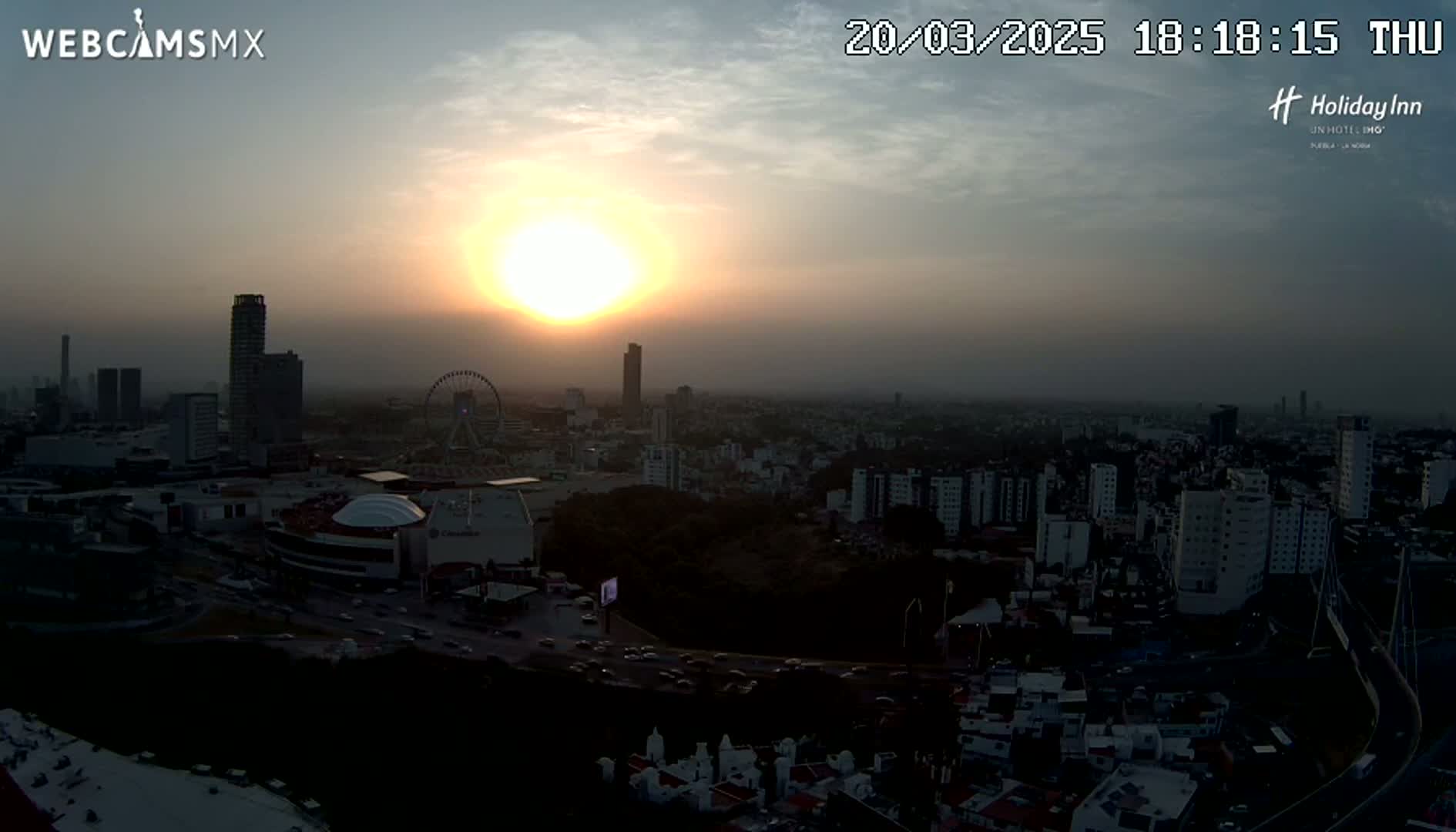 A hazy sunset illuminates a sprawling city, featuring several high-rise buildings, a Ferris wheel, and a network of roads.