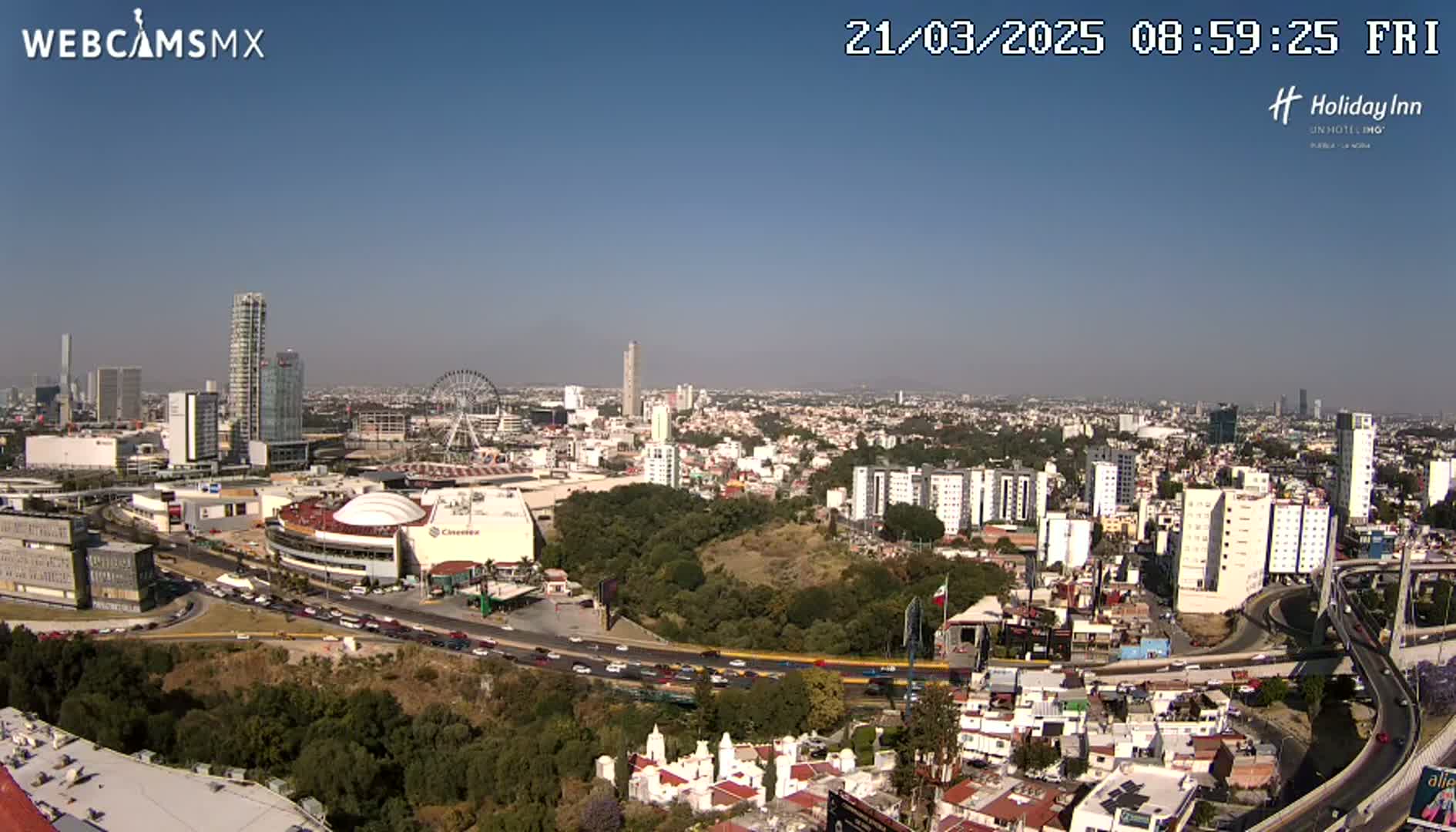 A panoramic view of a city on a clear, sunny day, showing a mix of high-rise buildings, residential areas, a Ferris wheel, and a large highway interchange.