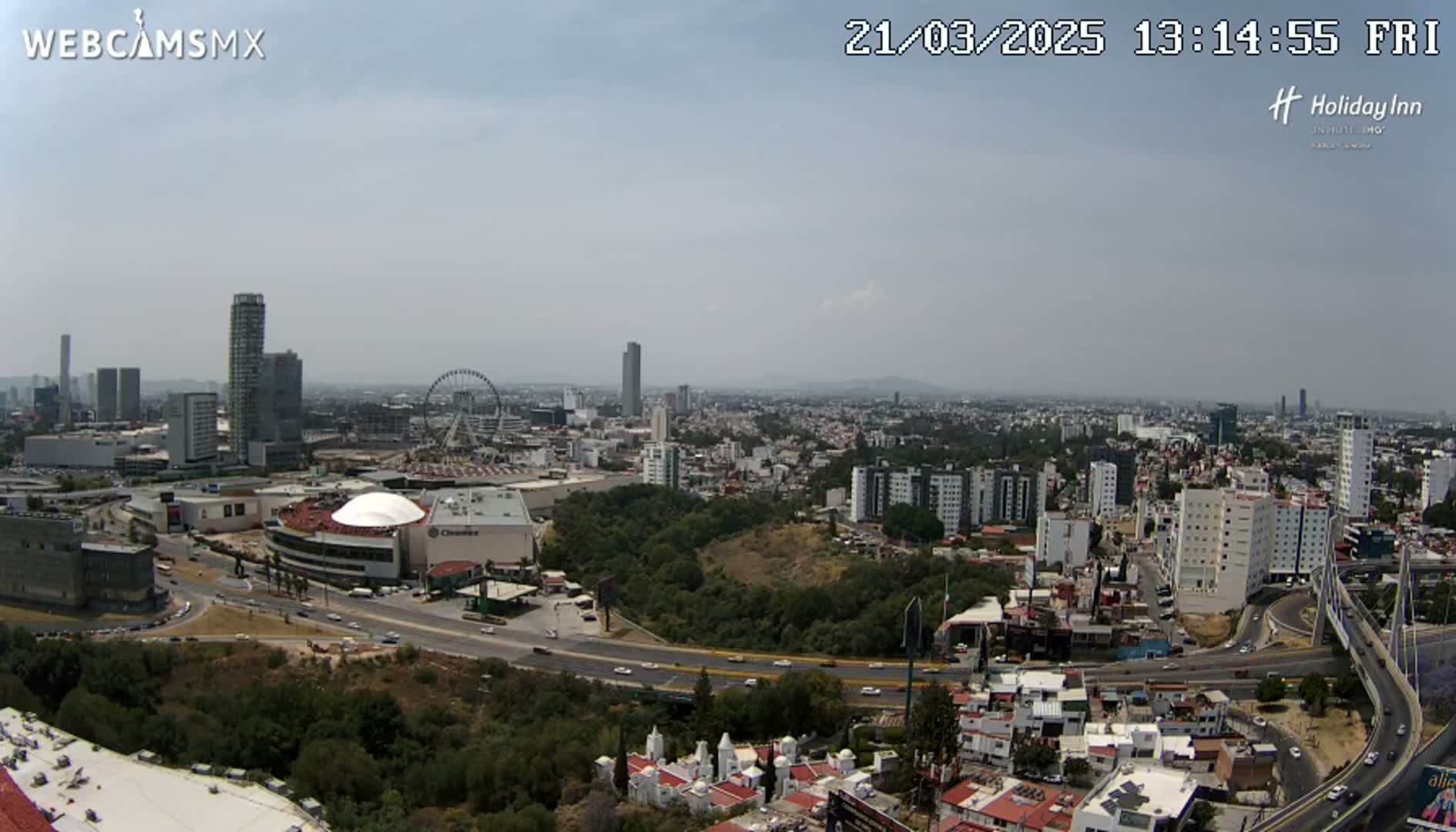 A hazy, sunny day reveals a sprawling city landscape with a large shopping mall, Ferris wheel, and highway system, surrounded by green hills and residential areas.