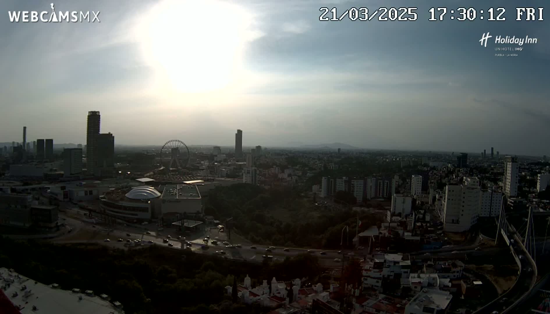 A hazy sunset illuminates a sprawling cityscape with high-rise buildings, a Ferris wheel, and a network of roads.