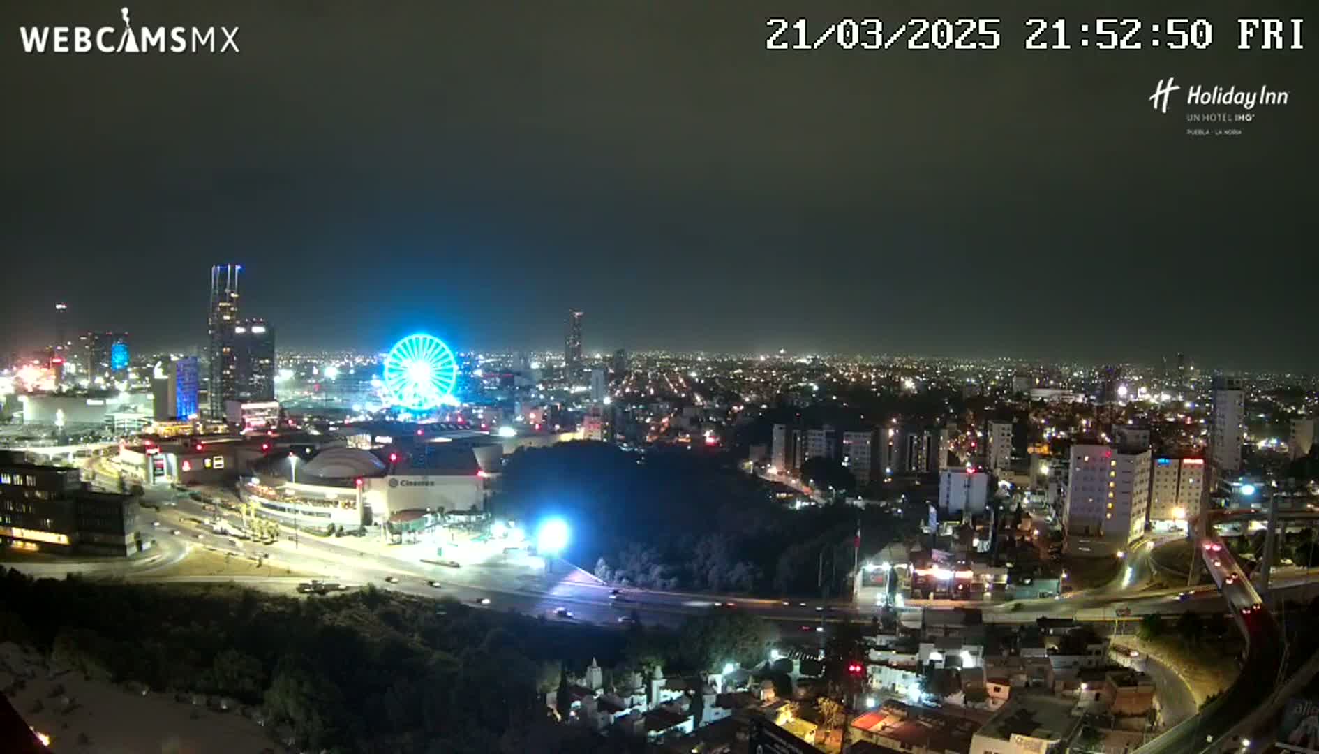 A nighttime aerial view of a city illuminated by lights, featuring a Ferris wheel and a large complex of buildings, under a clear, dark sky.