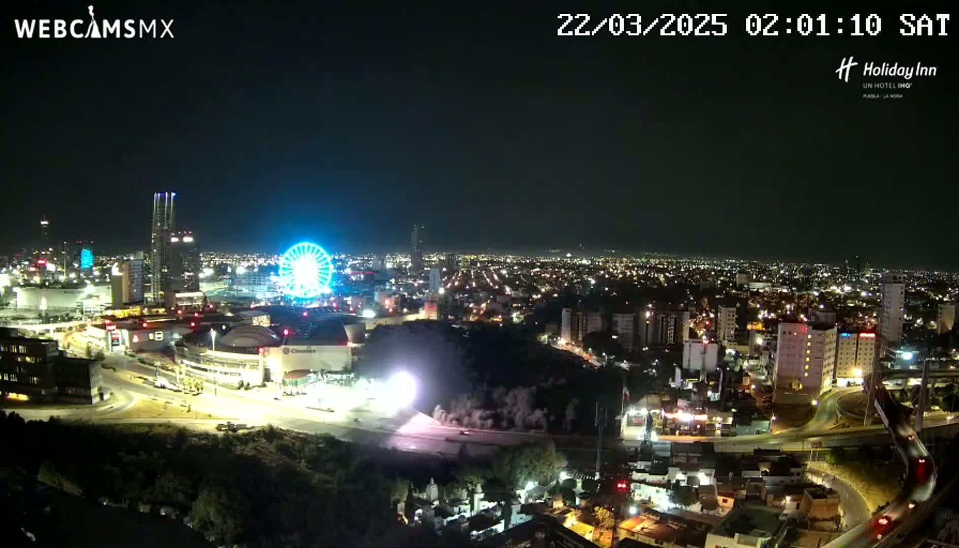 A nighttime, illuminated cityscape with a Ferris wheel, surrounding buildings, and roads visible under a clear sky.