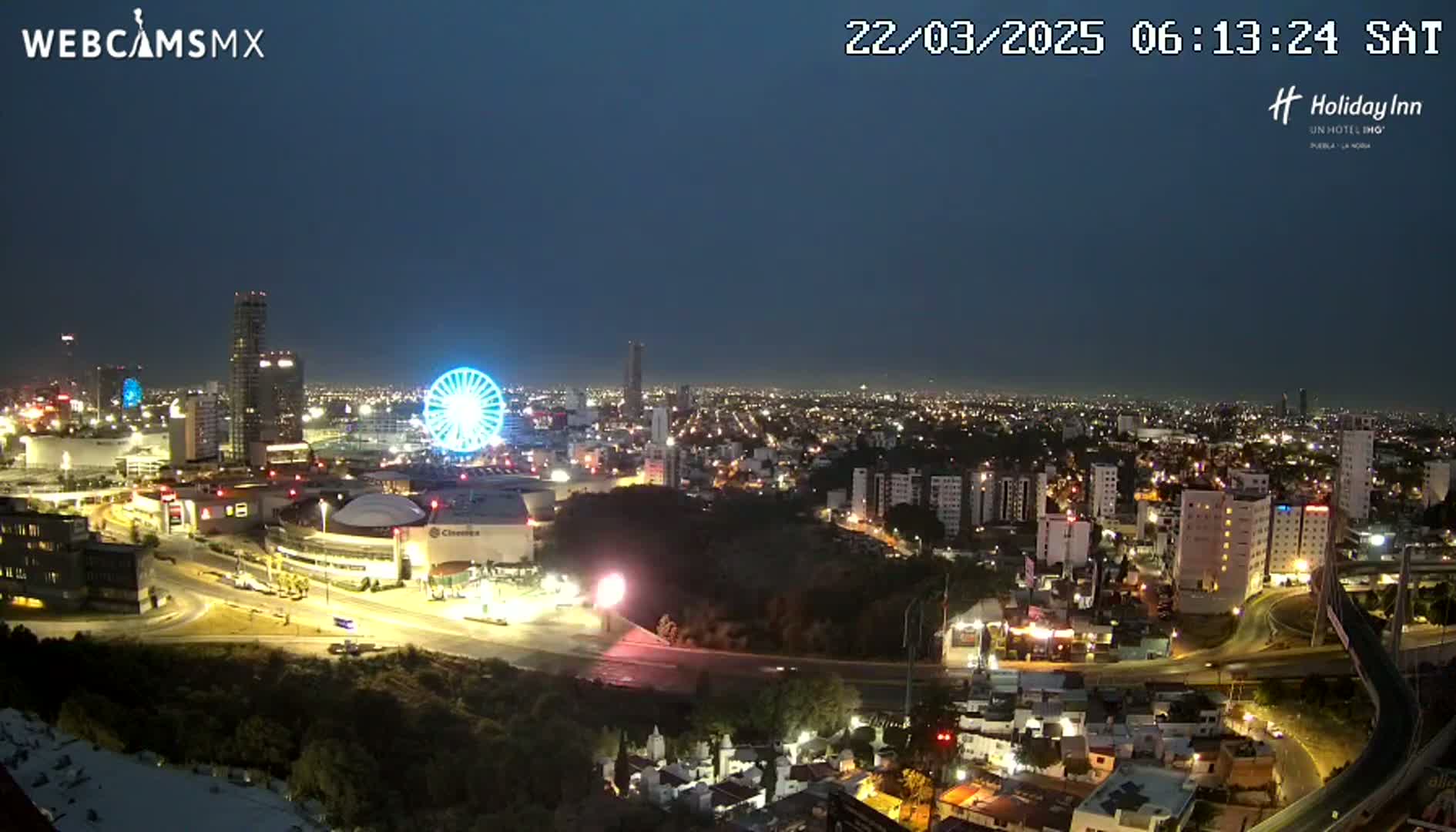 A nighttime view of a city with a Ferris wheel, illuminated buildings, and roads, under a dark blue sky.
