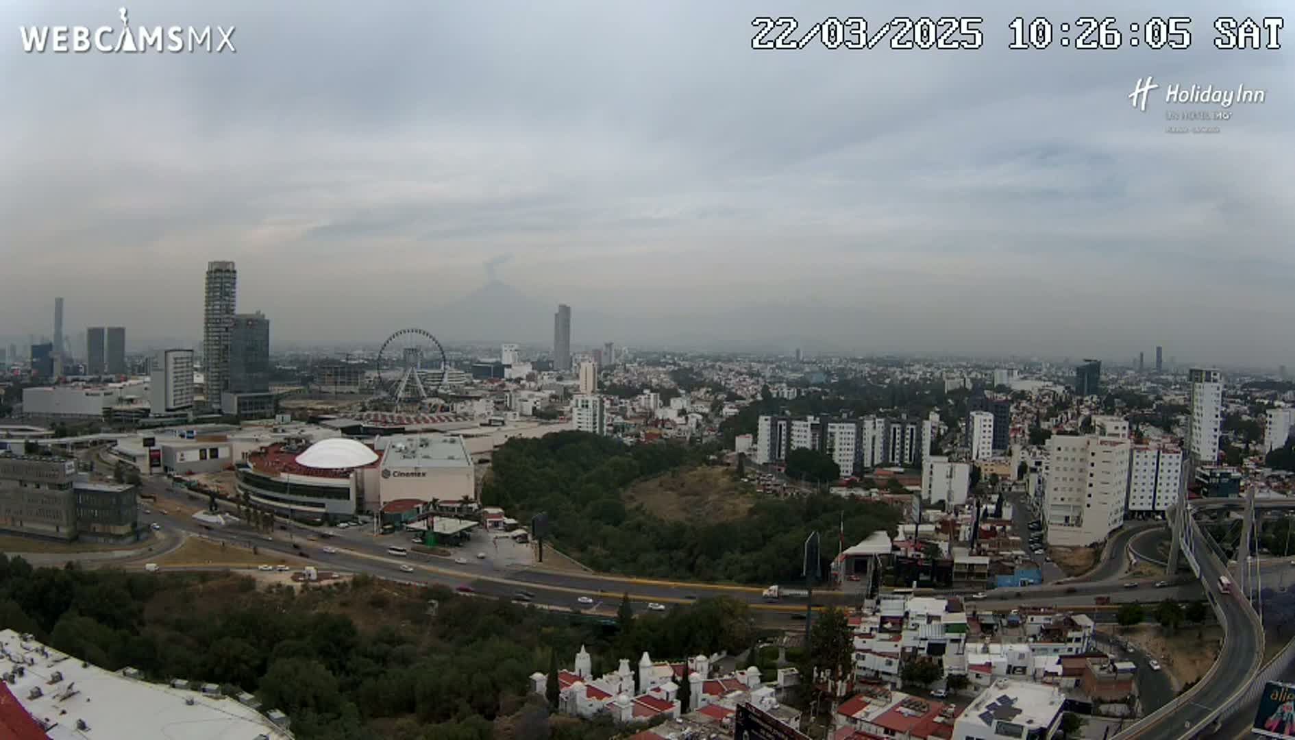 A hazy, overcast cityscape view shows a sprawling city with high-rise buildings, a Ferris wheel, and a distant volcano under a gray sky.