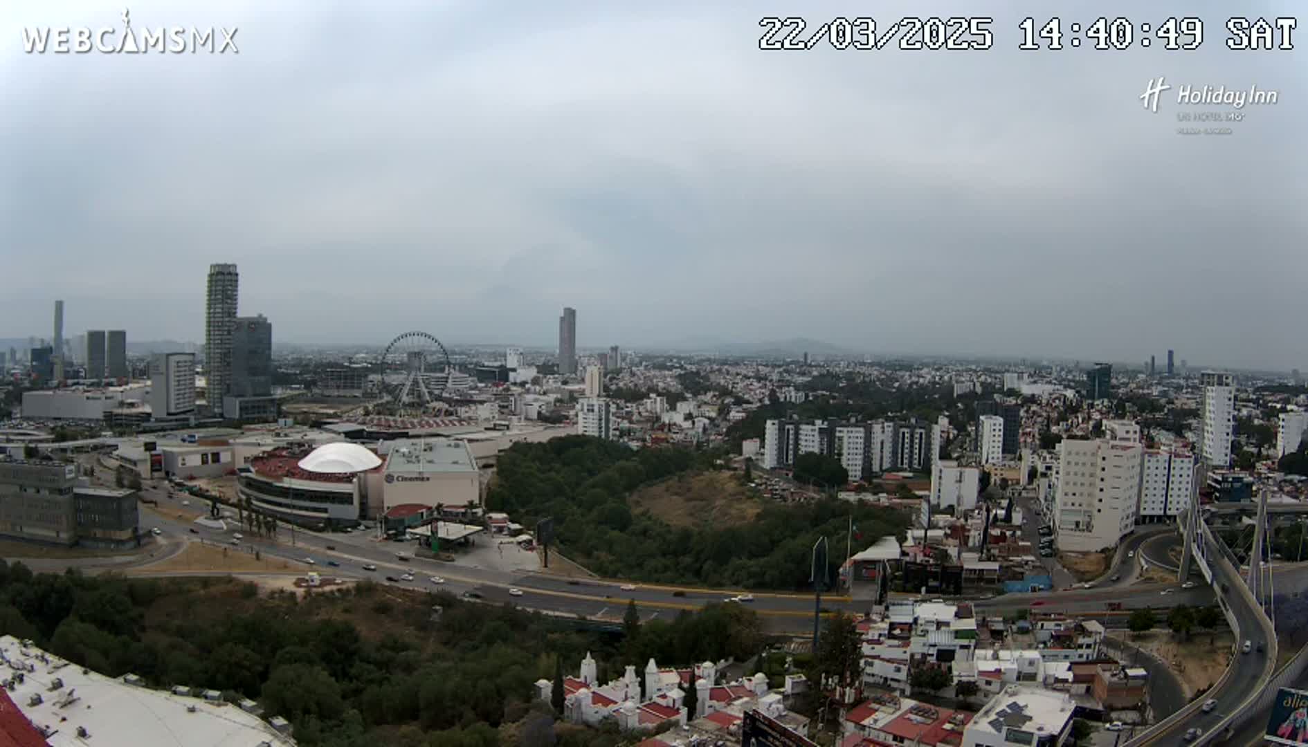 An aerial view of a city on an overcast day, showing a large shopping mall, a Ferris wheel, numerous buildings, and a highway system.