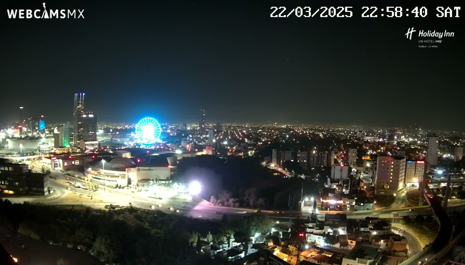 A nighttime cityscape with a Ferris wheel, illuminated buildings, and roadways, under a clear sky.