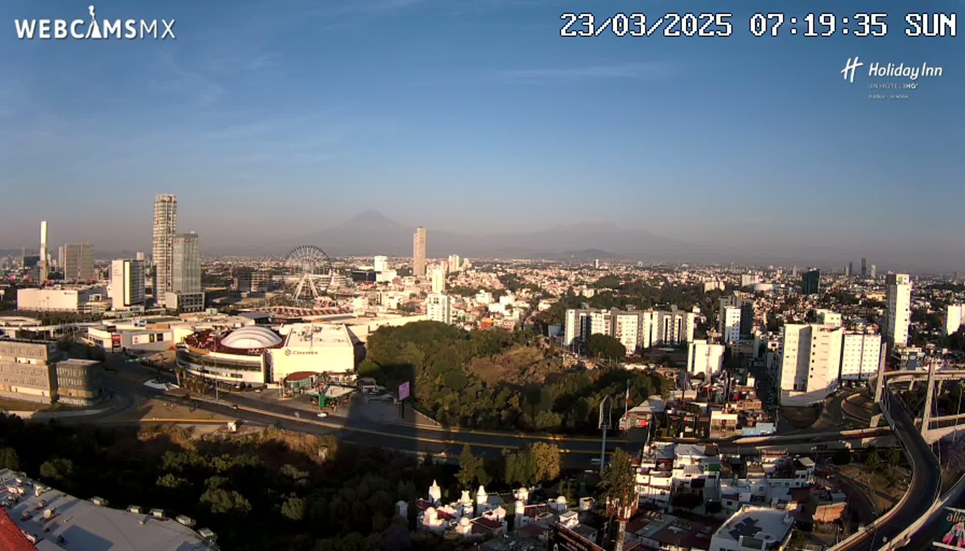 A sunny cityscape view shows a sprawling city with high-rise buildings, a Ferris wheel, and a distant mountain range under a clear blue sky.