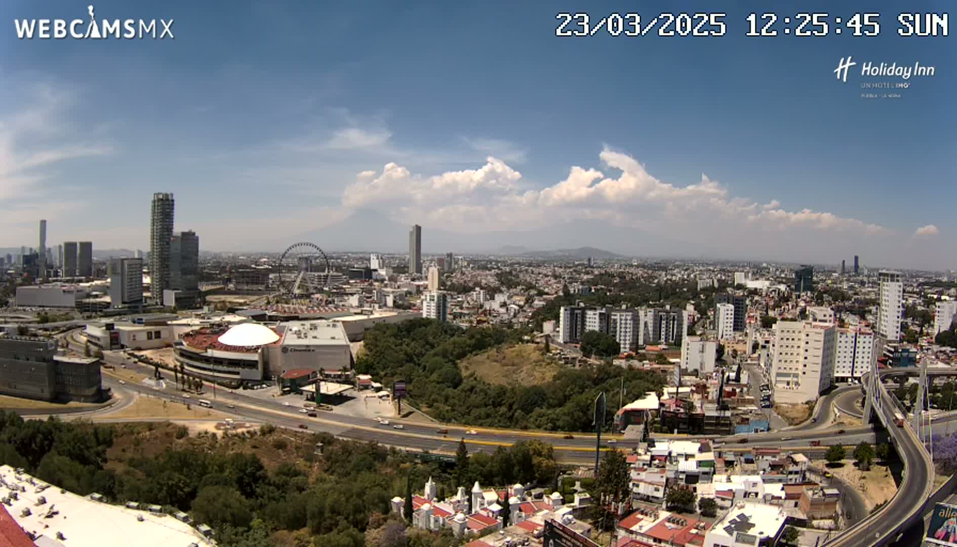 A sunny day reveals a sprawling cityscape with high-rise buildings, a Ferris wheel, and a large dome-shaped structure near a highway interchange, all surrounded by lush green areas.