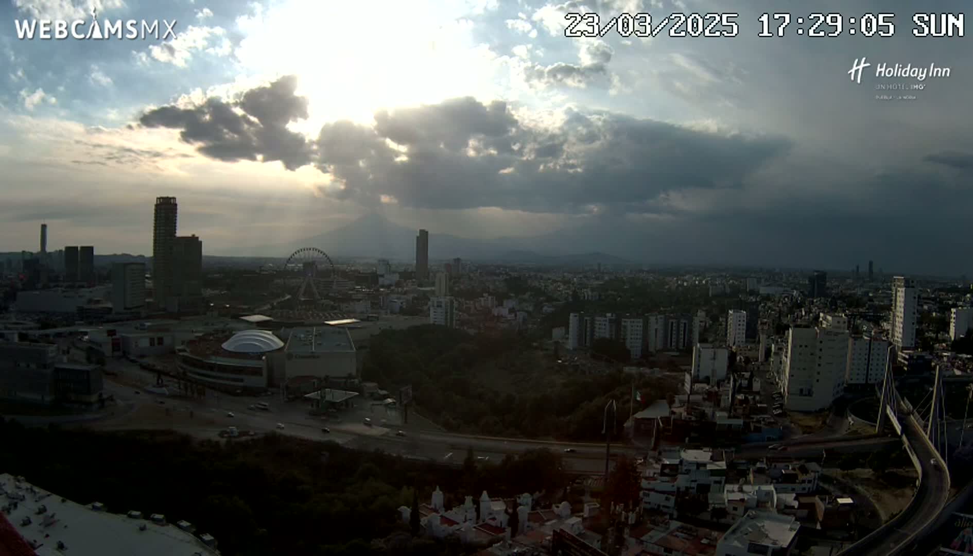 A partly cloudy sunset over a sprawling city with a Ferris wheel and mountains in the distance.