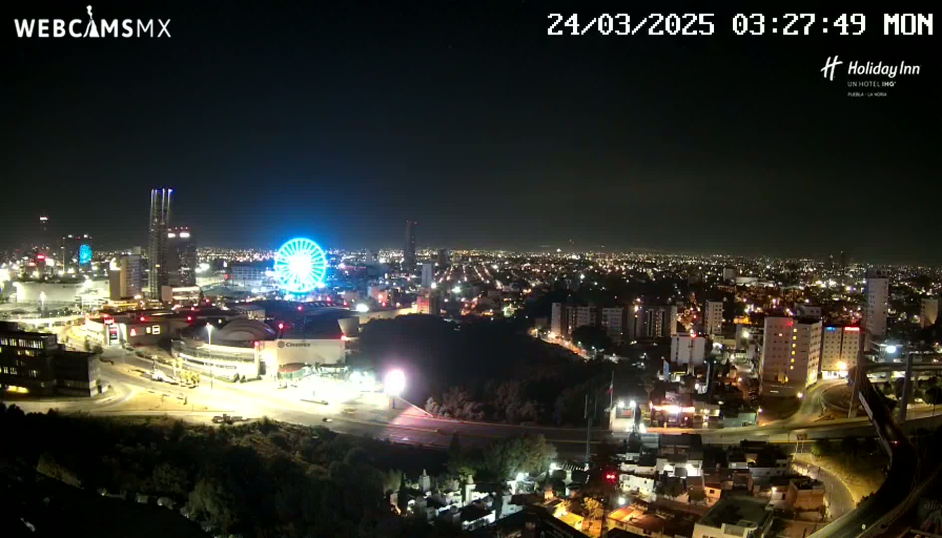 A nighttime, aerial view of a city with a Ferris wheel and illuminated buildings, under clear skies.