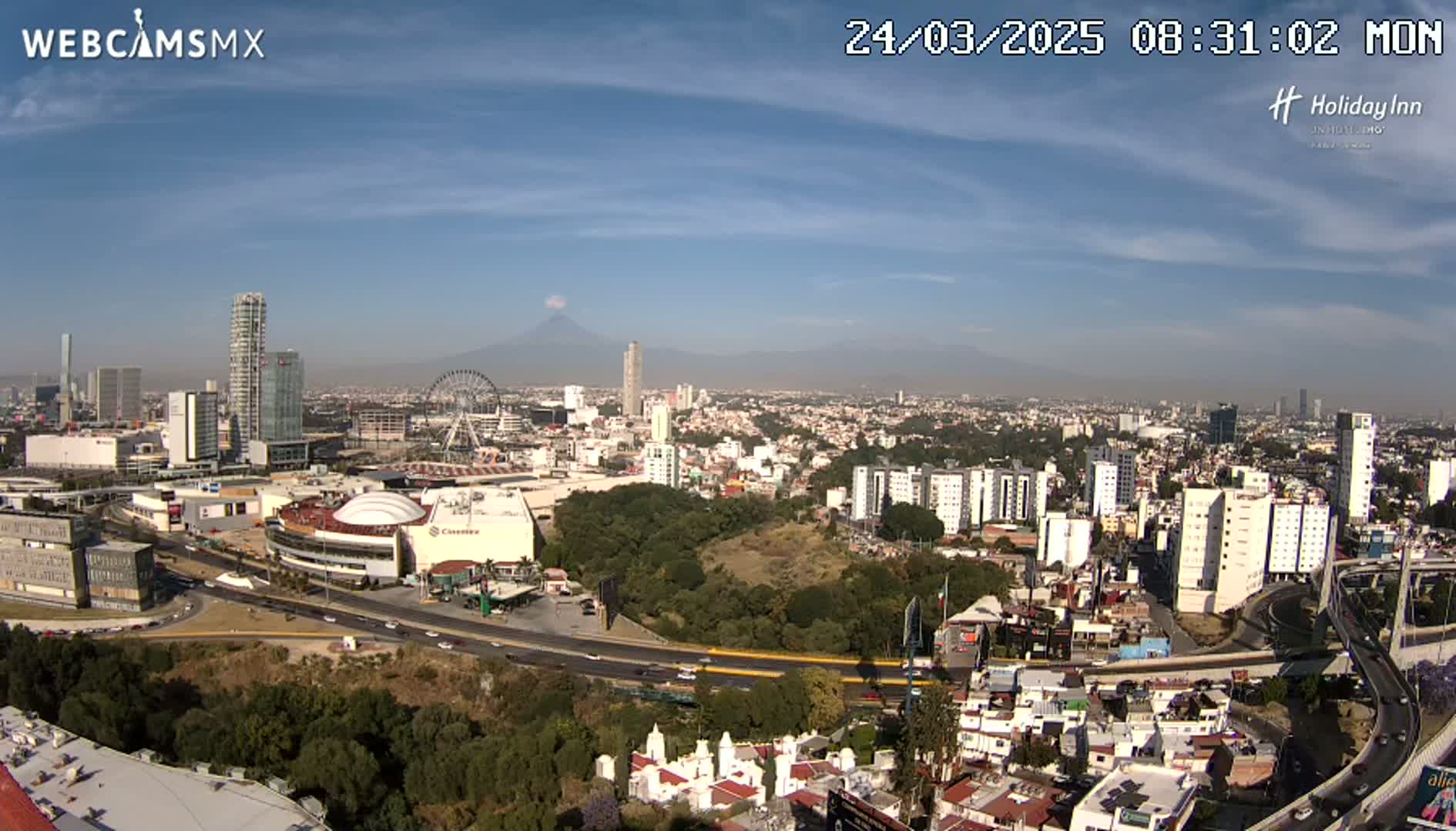A sprawling cityscape under a mostly clear, sunny sky, featuring a Ferris wheel, numerous buildings, and a distant volcano in the background.
