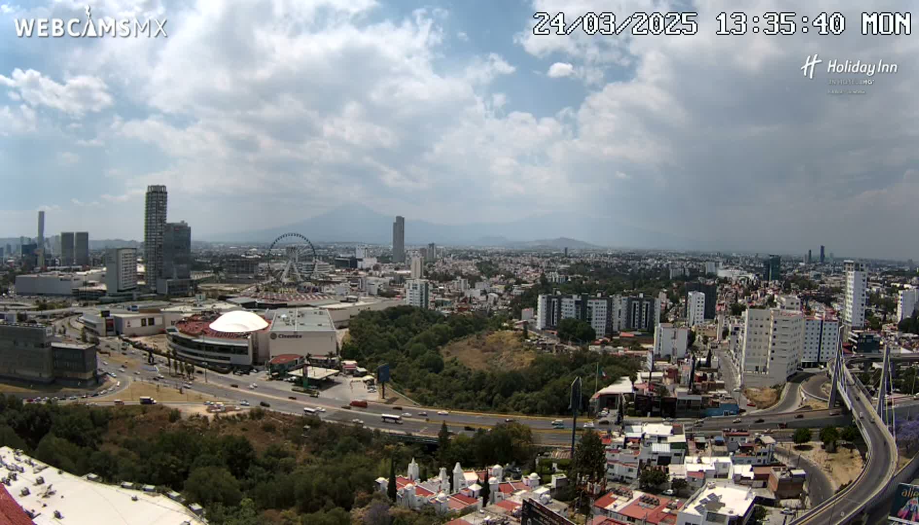 A partly cloudy daytime view of a sprawling city with a Ferris wheel, several high-rise buildings, and a large highway system, all set against the backdrop of distant mountains.