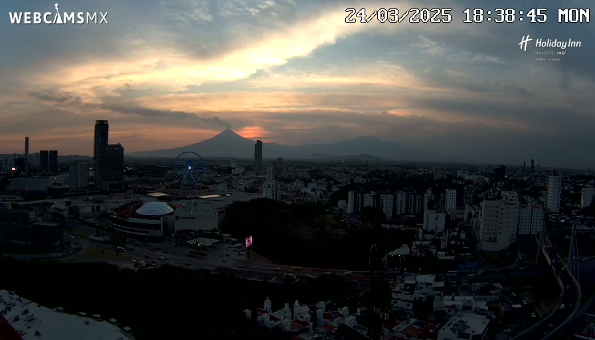 A sunset over a city with a Ferris wheel and a volcano visible in the distance, under partly cloudy skies.