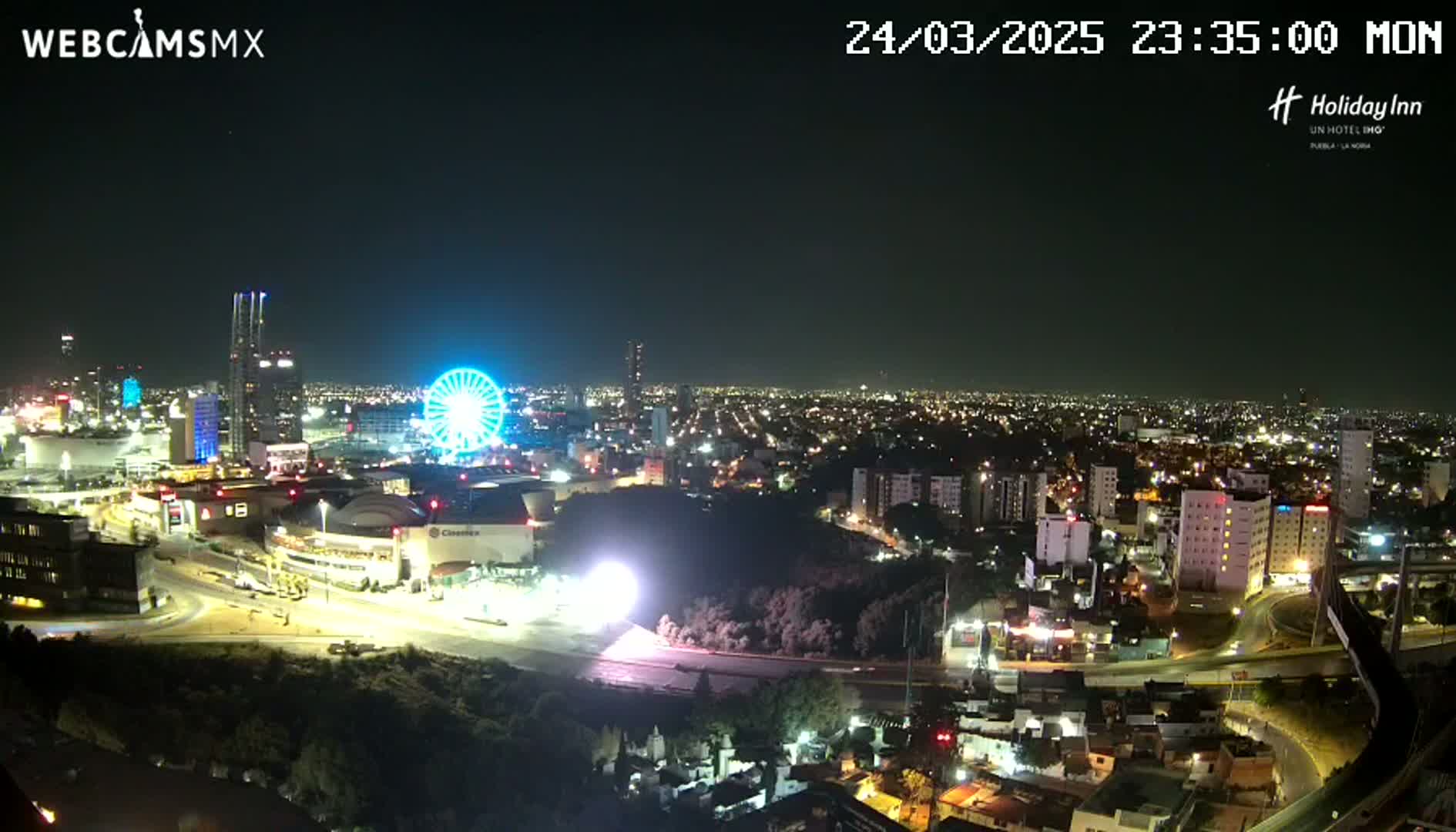A nighttime, aerial view of a city with a Ferris wheel illuminated, surrounded by buildings and roads.