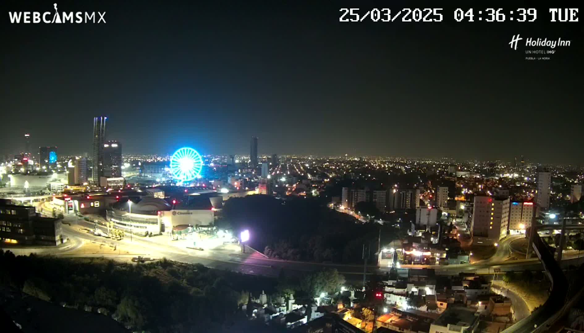 A nighttime, aerial view of a city illuminated by streetlights, featuring a Ferris wheel and several tall buildings, under a clear sky.
