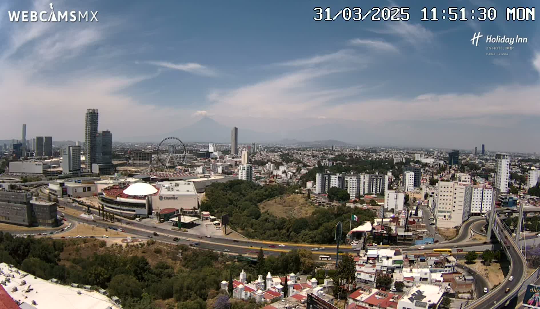 A sprawling city landscape under a mostly clear, sunny sky, featuring a Ferris wheel, several skyscrapers, a volcano in the distance, and a large highway system.
