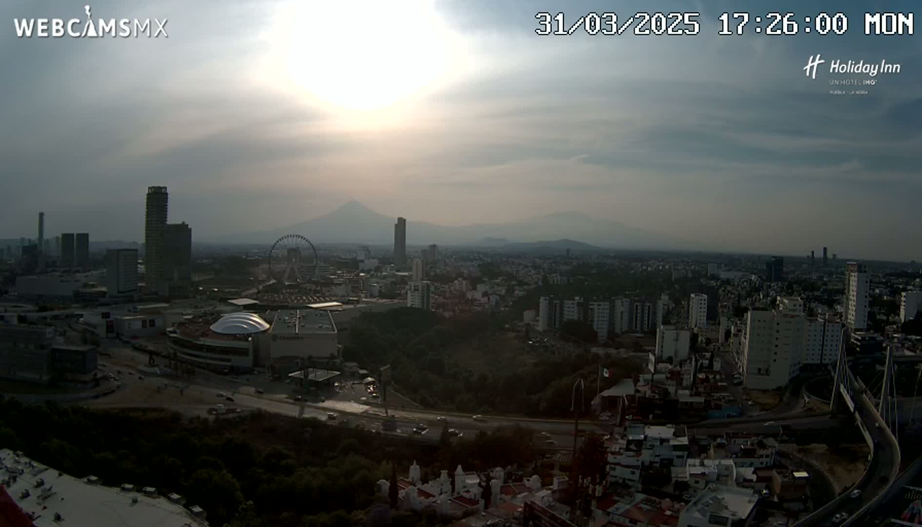 A hazy sunset illuminates a sprawling city landscape featuring skyscrapers, a Ferris wheel, and distant mountains.