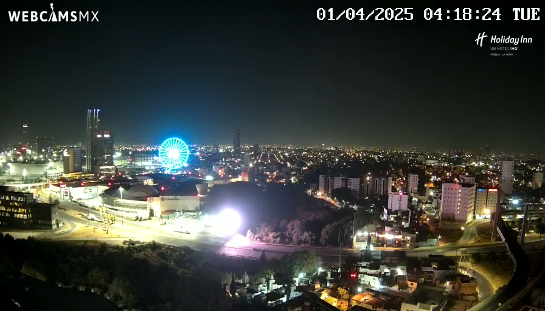 A nighttime aerial view of a city with a Ferris wheel and brightly lit buildings, under a clear sky.