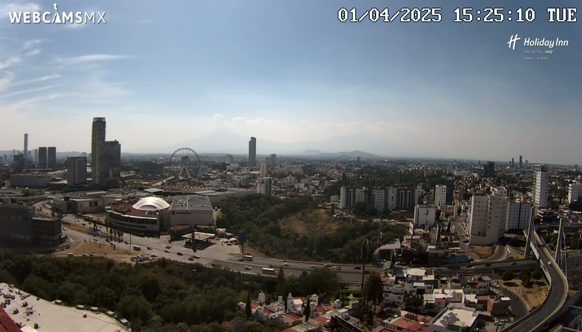 A high-angle, long shot on a clear, sunny day shows a sprawling city with several high-rise buildings, a Ferris wheel, and a large shopping mall, all surrounded by green hills and a network of roads and bridges.