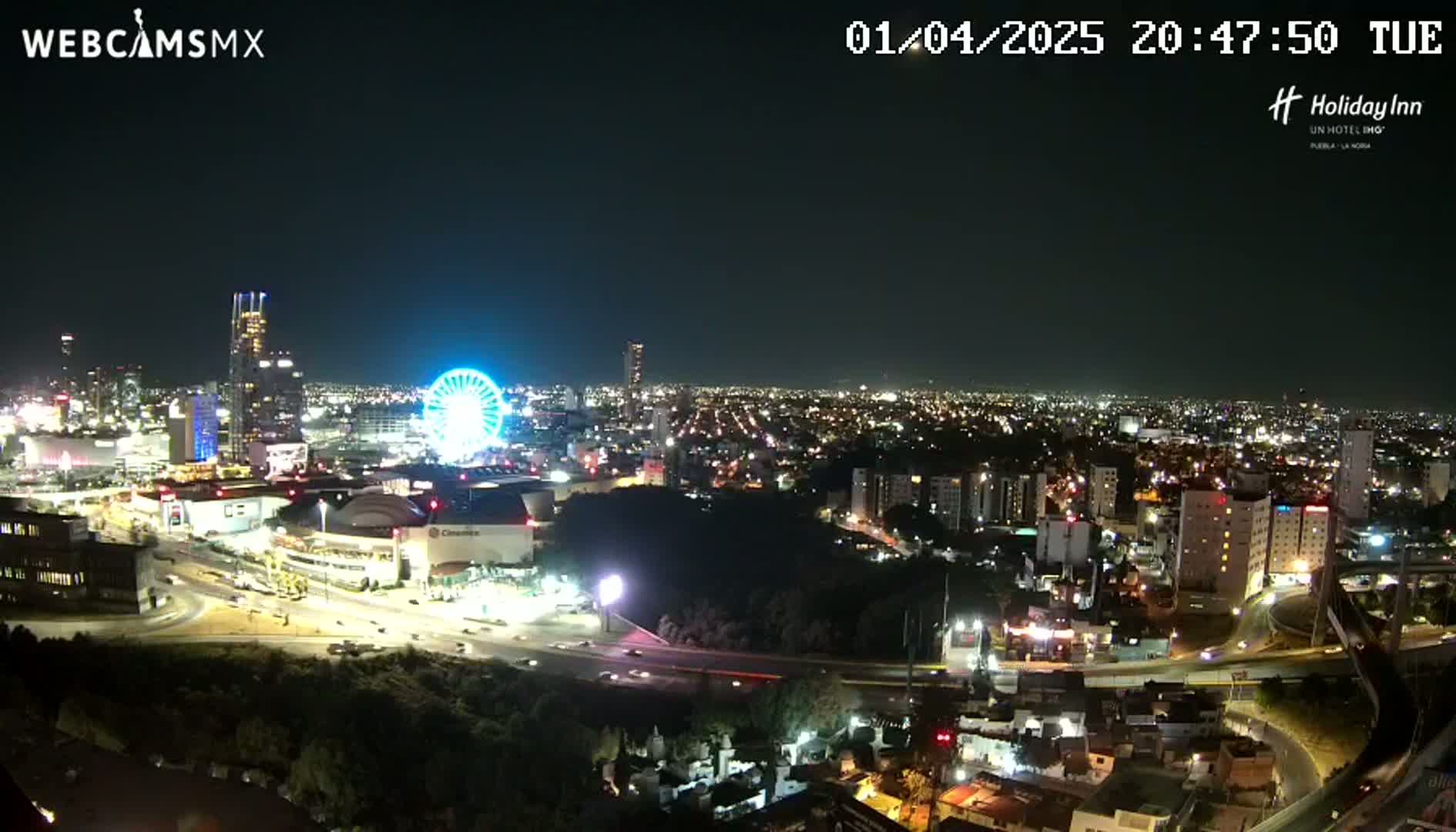 A nighttime, aerial view of a city with a Ferris wheel and illuminated buildings, under a clear, dark sky.